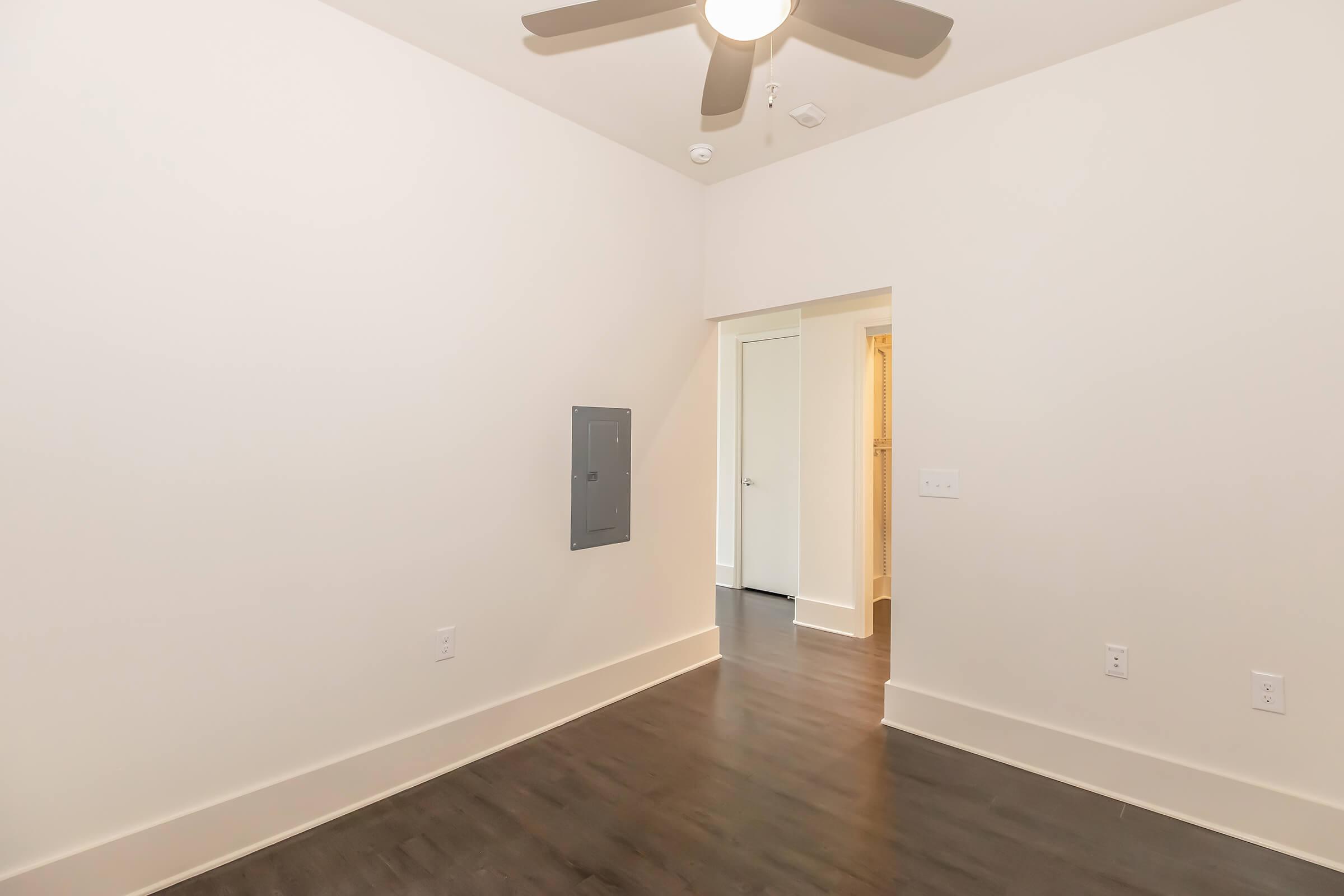Empty room with light-colored walls and dark flooring. A ceiling fan is installed, and there is a small electrical panel on one wall. An open doorway leads to another area, suggesting a spacious layout. The overall atmosphere is bright and minimalistic.