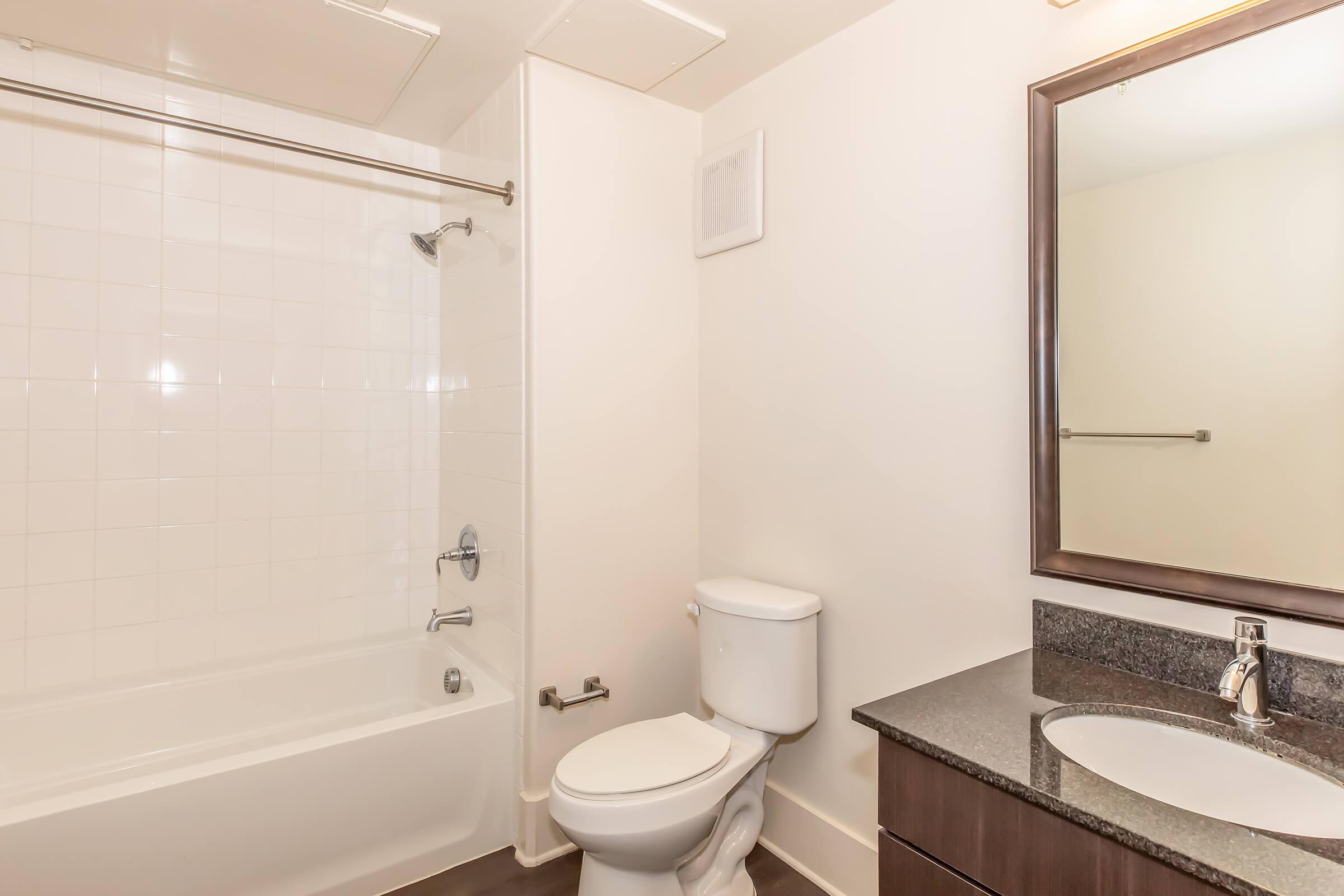 A clean, modern bathroom featuring a bathtub with a shower, a white toilet, and a dark granite countertop with a sink. A mirror is mounted above the sink, and the walls are tiled in white, creating a bright and spacious look.