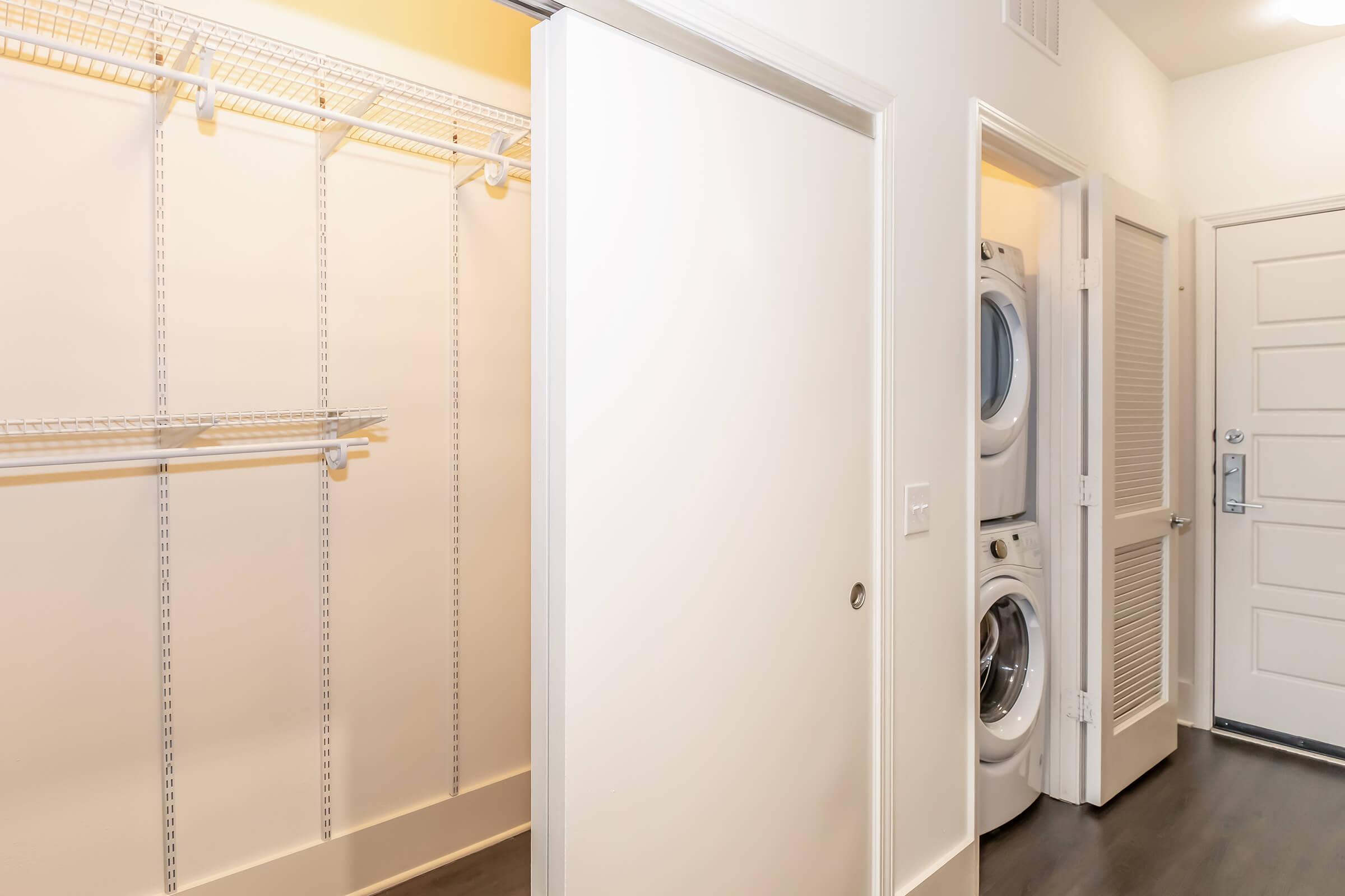 Interior view of a laundry area featuring a closet with wire shelving on one side and a stacked washer and dryer on the other. The space has modern white walls and a door leading to another room, creating a functional and organized utility space.