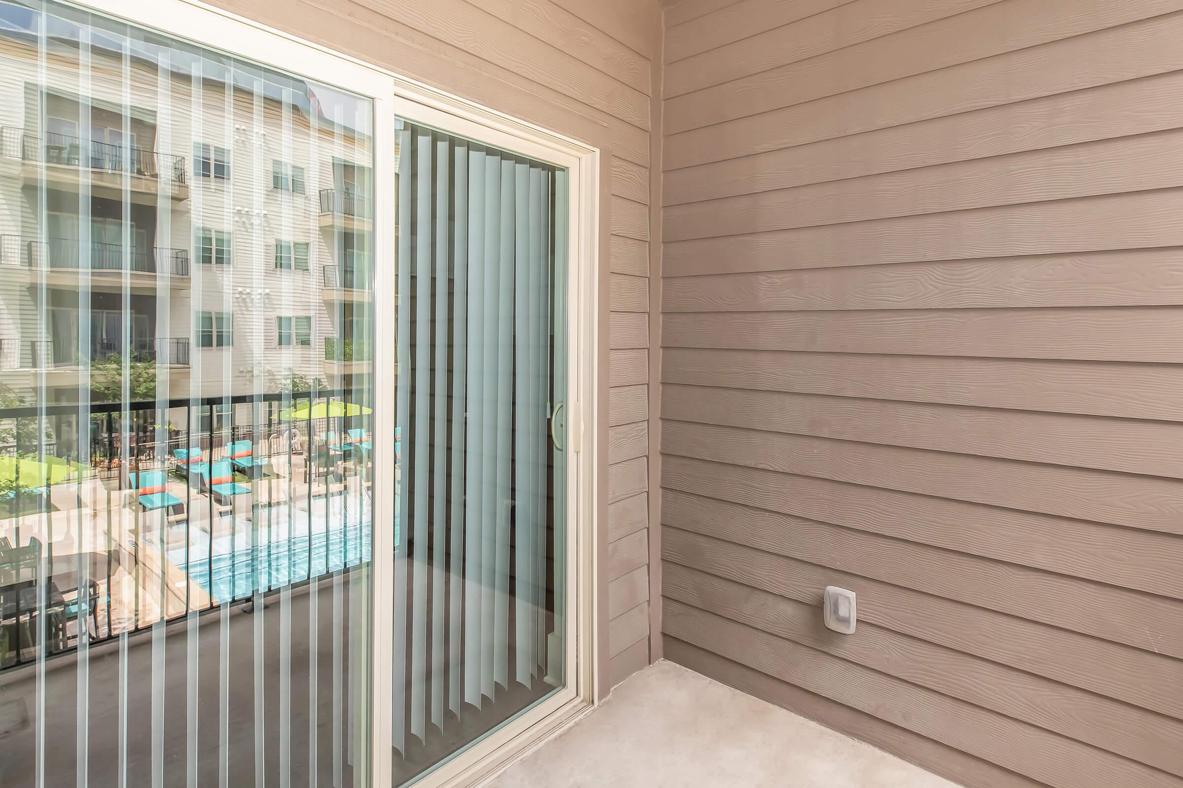 A view of a balcony with a sliding glass door featuring vertical blinds. Visible through the door is a swimming pool area with lounge chairs and umbrellas, surrounded by an apartment complex with multiple stories. The balcony has beige siding and a clean, empty floor.
