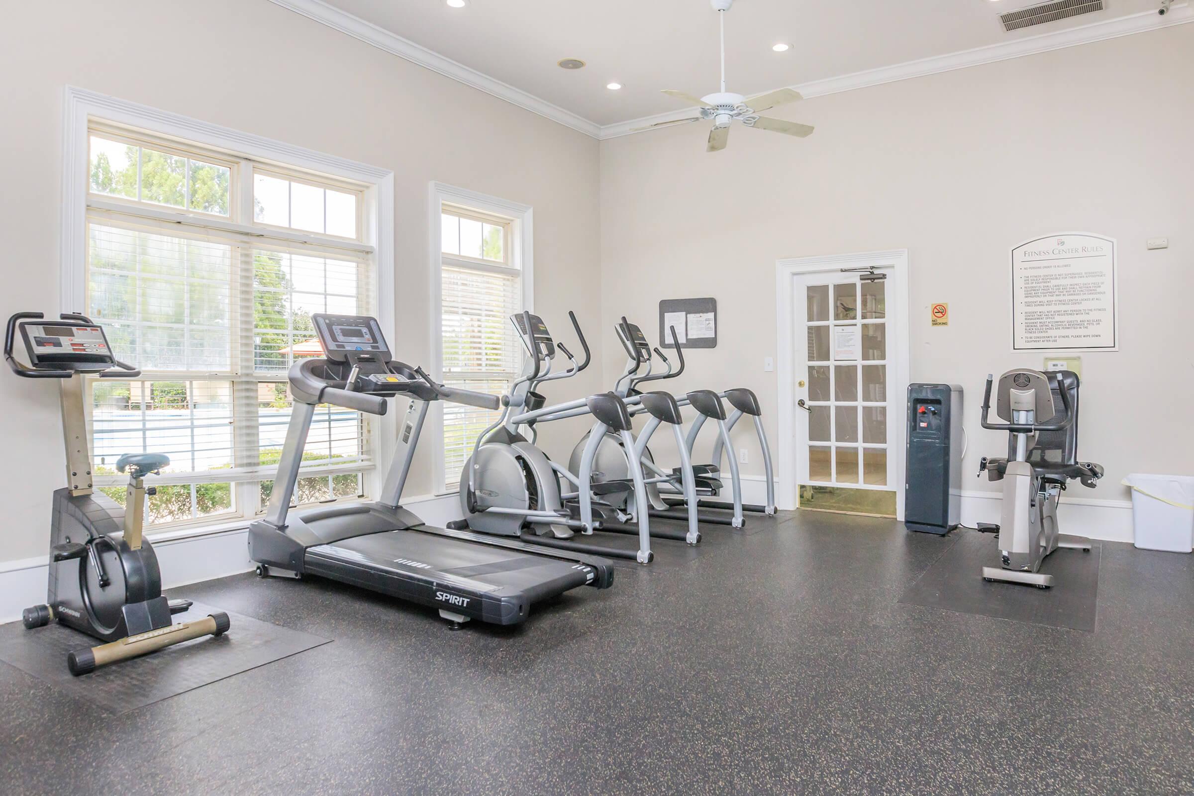 A fitness room featuring various gym equipment including treadmills, elliptical machines, and a stationary bike. Large windows provide natural light, and there is a water cooler and a sign on the wall with information. The flooring is dark and rubberized for safety.