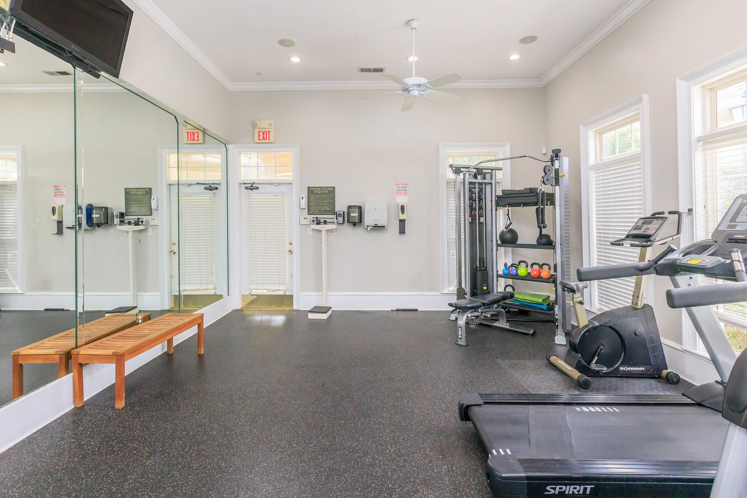 A well-lit gym area featuring various exercise equipment including treadmills, a multi-station weight machine, and free weights. There’s a wooden bench against the wall and large mirrors reflecting the space. Two exit doors are visible, with fitness accessories organized neatly.