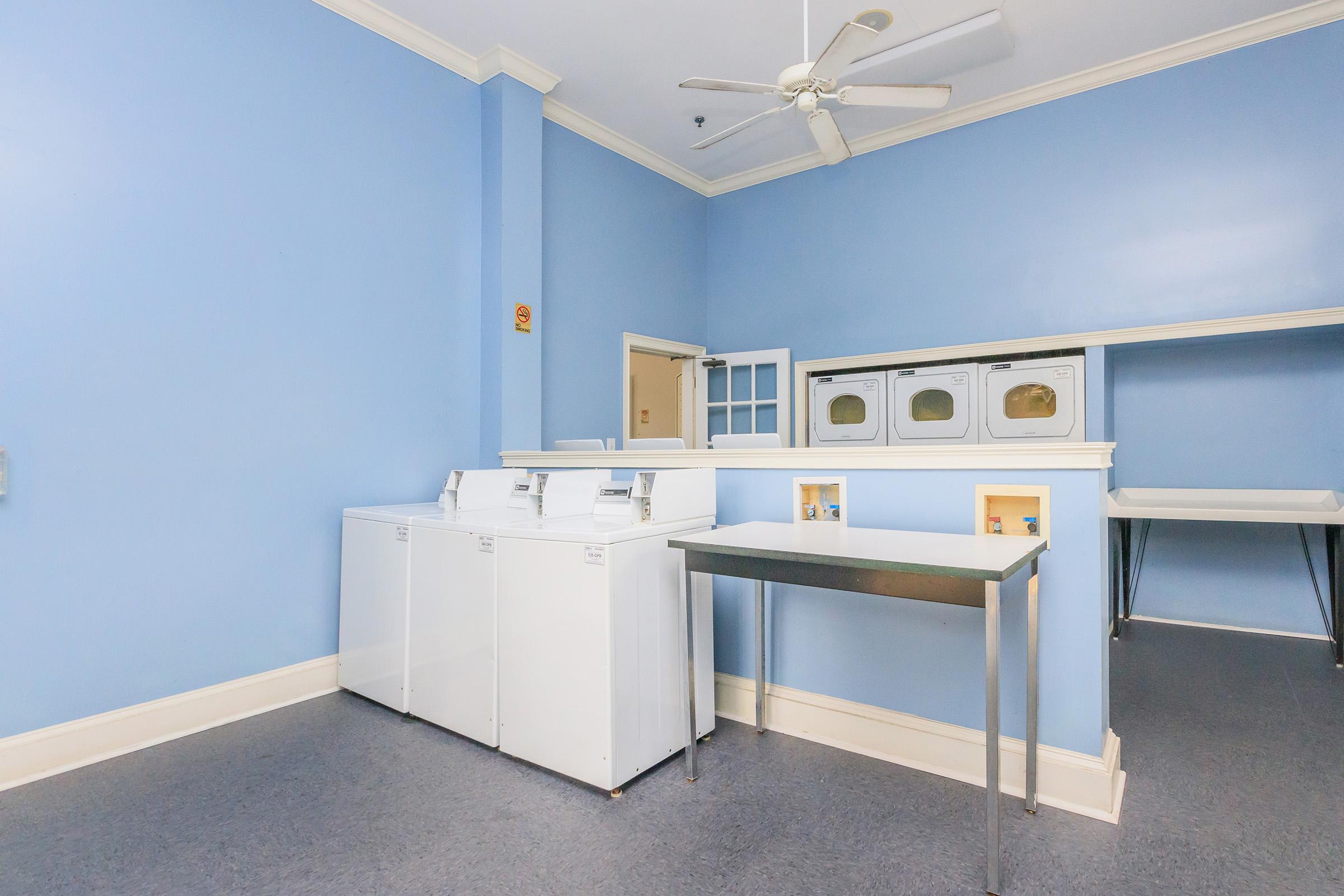A bright and clean laundry room featuring two white washing machines and several stacked dryers against a light blue wall. There's a stainless steel folding table on the left and a ceiling fan above, creating a spacious and organized atmosphere.