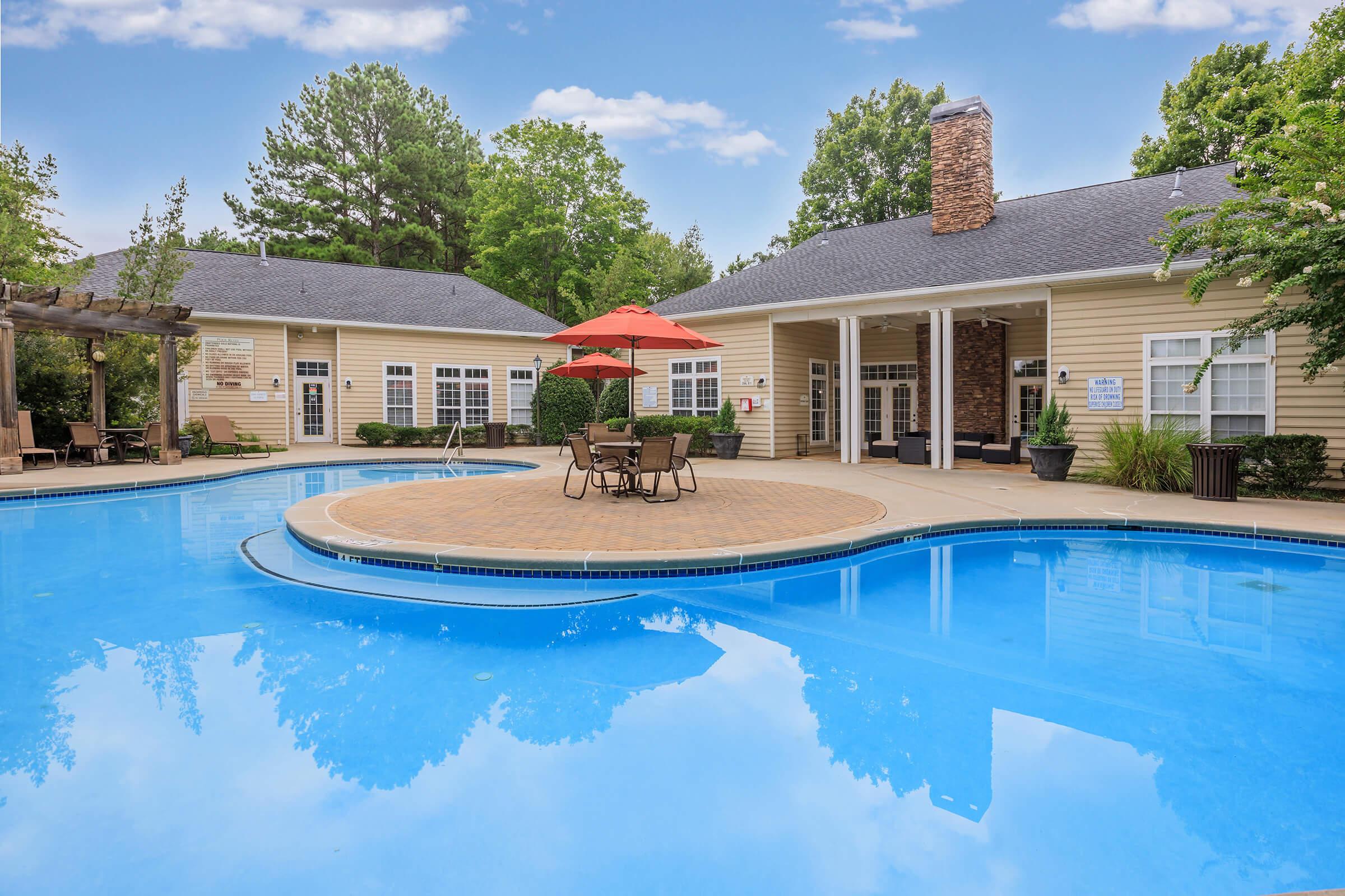 A clear blue swimming pool surrounded by lounge chairs and a bright orange umbrella. In the background, there is a clubhouse with large windows and a stone chimney, complemented by lush greenery and trees. The setting is inviting and ideal for relaxation or socializing.
