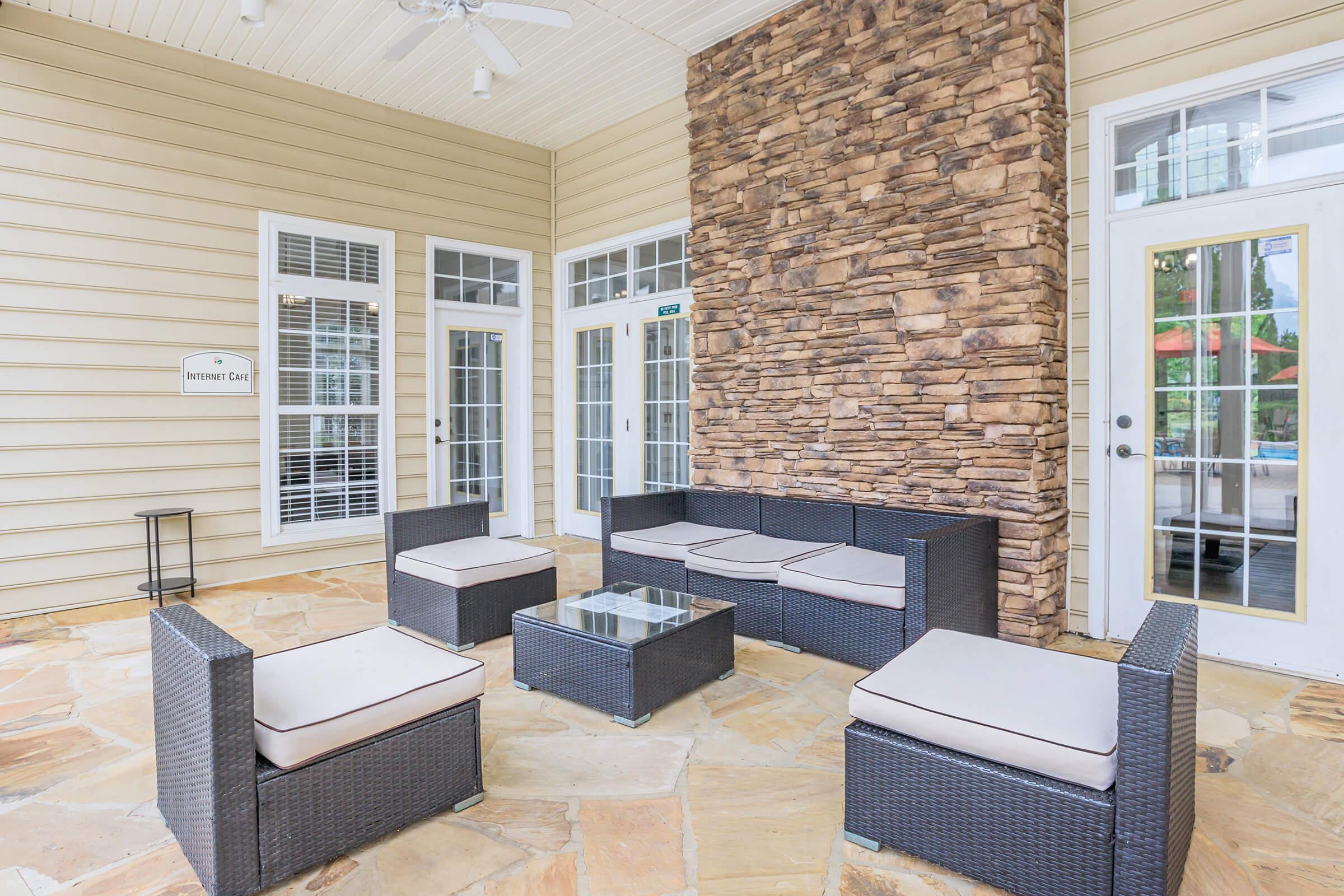 Seating area with modern wicker furniture, including a coffee table and cushions, set against a decorative stone wall. The space features large windows and doors leading to an outdoor area, with a neutral color palette and a cozy ambiance.