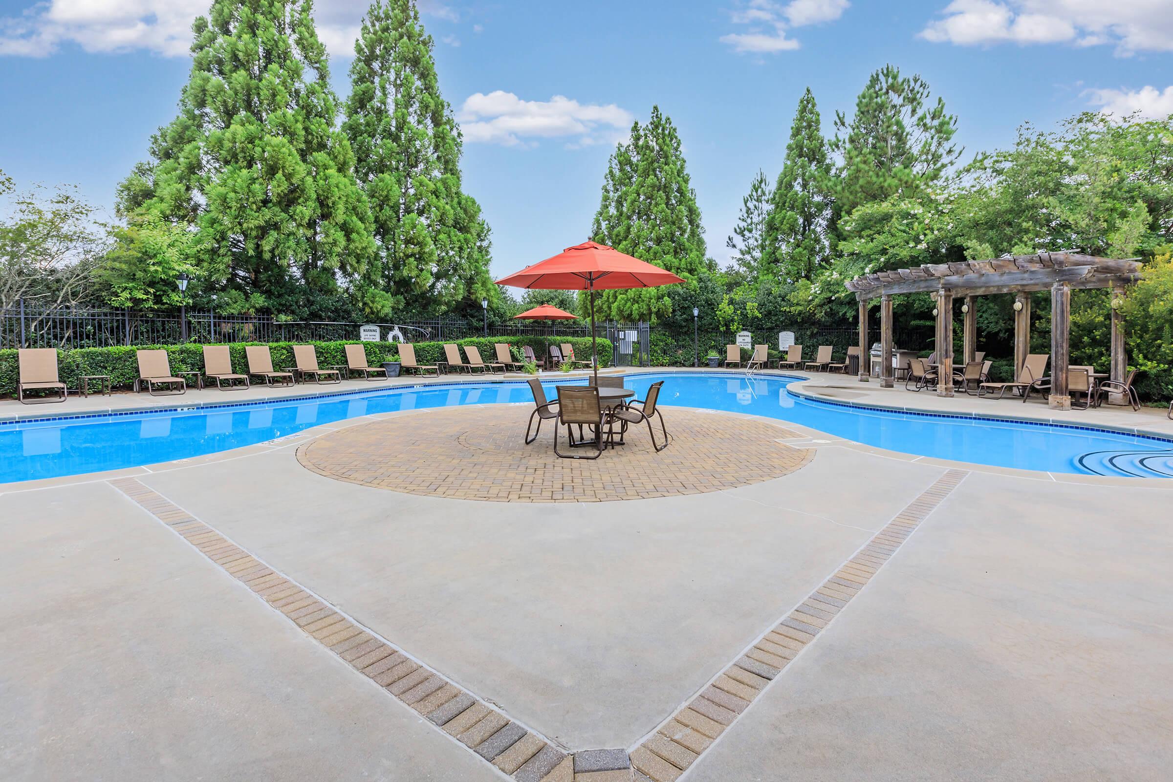 A well-maintained swimming pool surrounded by lounge chairs and green trees. In the center, there’s a circular seating area with a table and chairs shaded by a red umbrella. A pergola is visible to the right, creating a relaxing outdoor atmosphere against a clear blue sky.