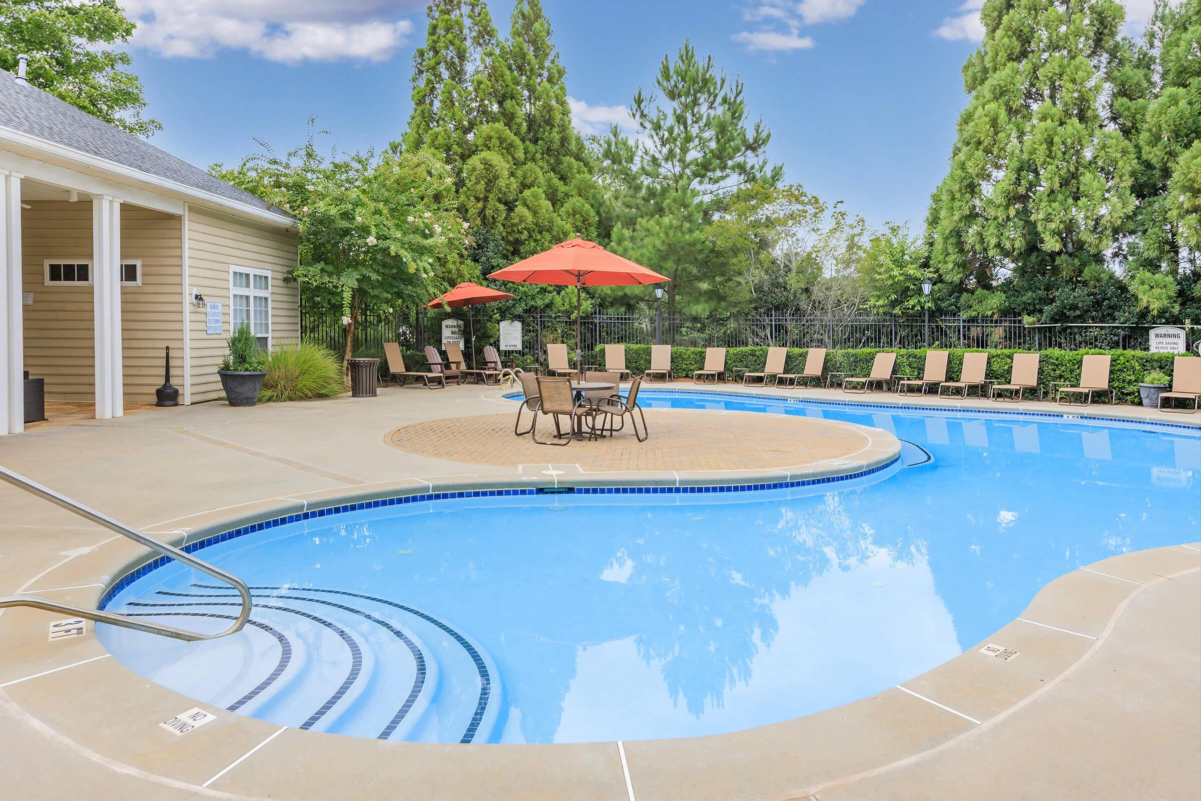 A sunny outdoor pool area featuring a circular pool with a blue surface, surrounded by tan lounge chairs and orange umbrellas. Lush green trees provide a backdrop, and a beige building is visible to the side. The scene conveys a relaxing and inviting atmosphere.