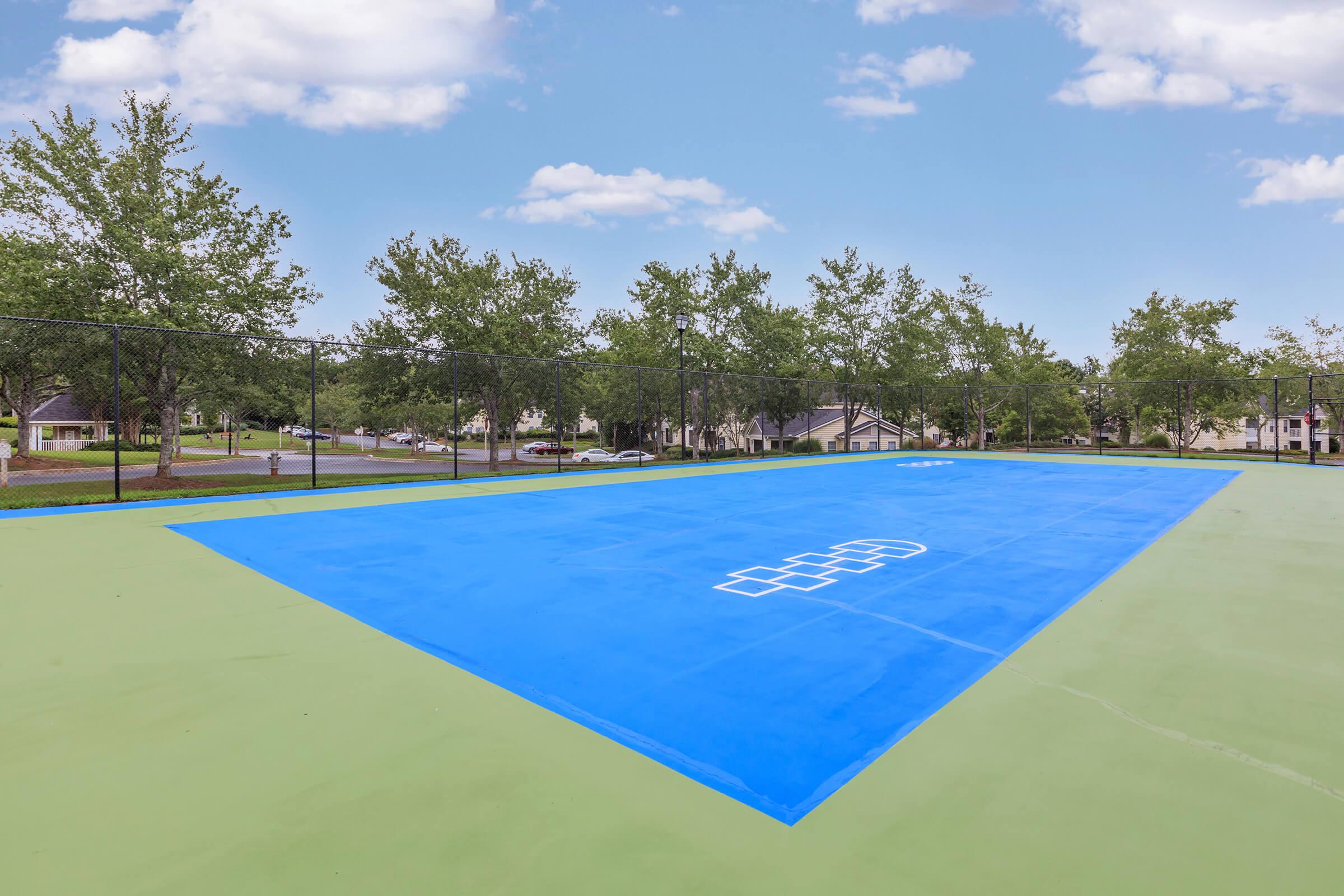 A brightly colored outdoor basketball court featuring a blue playing area and green surrounding surface. The court is surrounded by trees and residential buildings in the background, under a clear blue sky with white clouds.