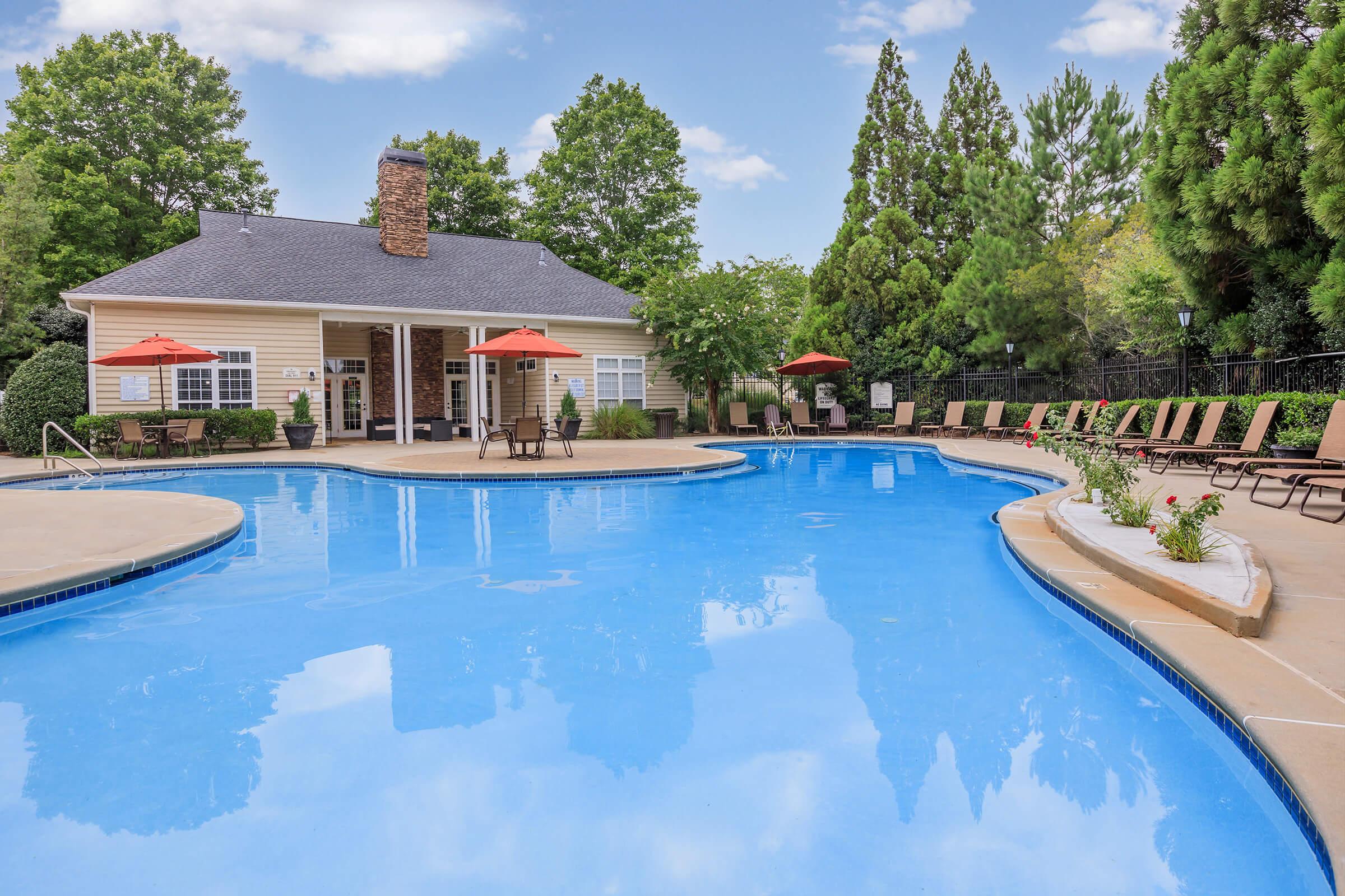 A serene swimming pool surrounded by lush greenery, with lounge chairs and umbrellas. In the background, there is a charming building with large windows, offering a welcoming atmosphere. The sky is clear and blue, creating a perfect setting for relaxation.