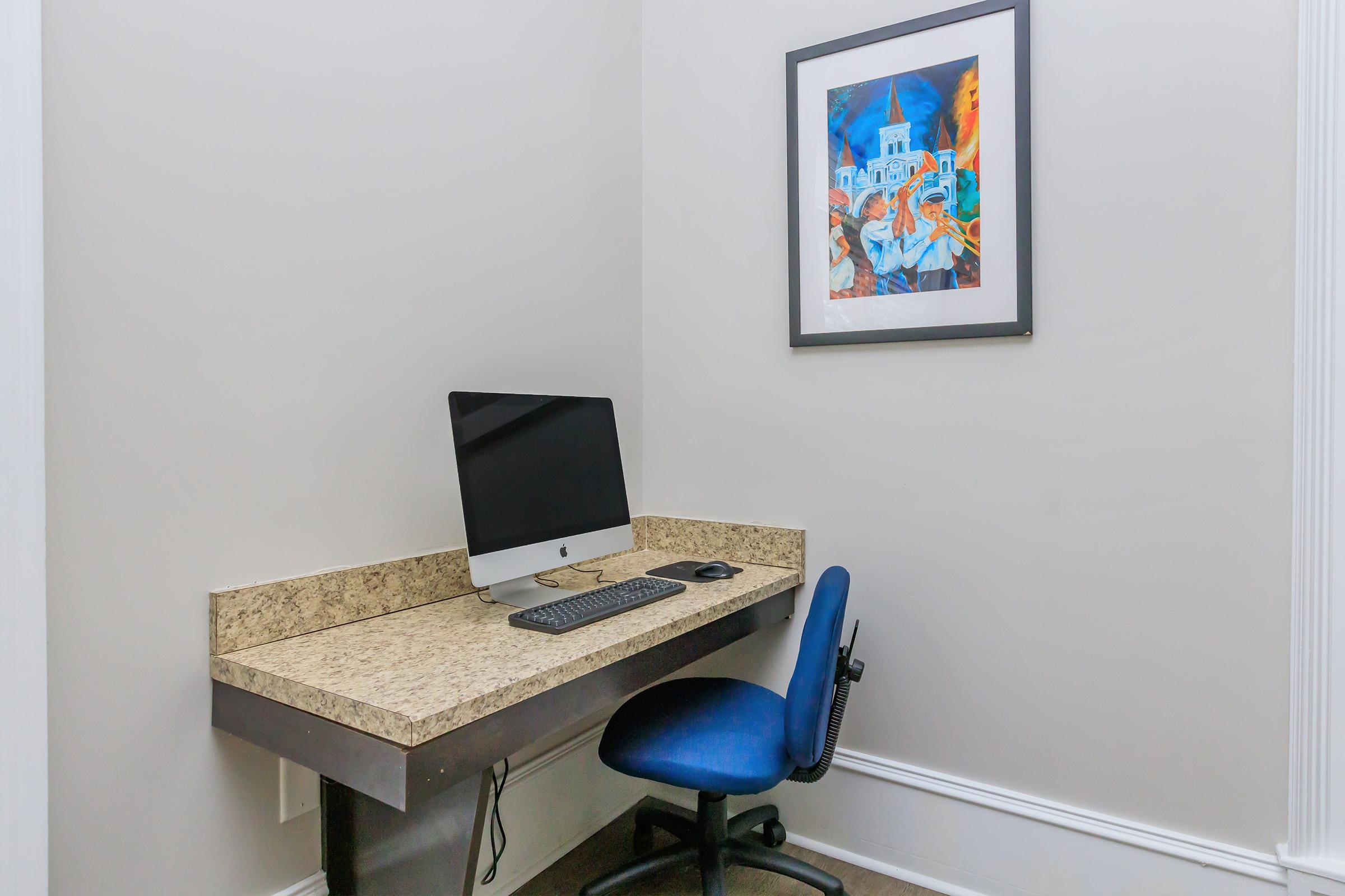 A minimalist workspace featuring a granite desk with a silver iMac computer, keyboard, and mouse. An office chair is positioned at the desk. The wall is painted a light color, and a colorful framed artwork is displayed above the desk, adding a touch of creativity to the space.