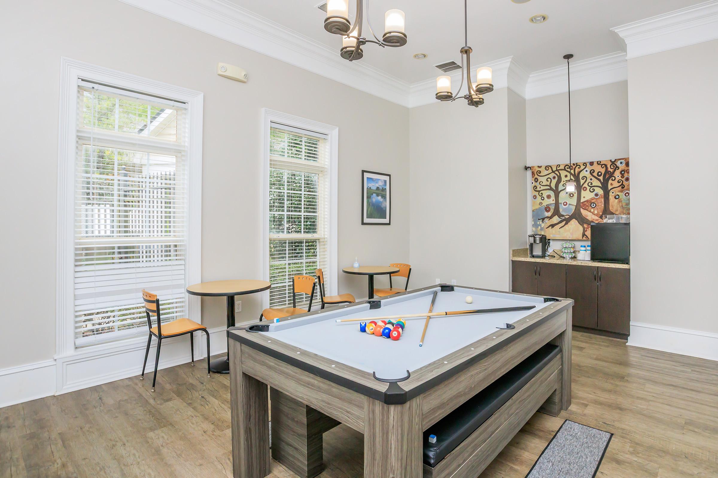 A well-lit game room featuring a pool table with colorful balls, a modern chandelier, and two small tables with chairs. The walls are painted a neutral color, and windows allow natural light. An artistic wall decoration and a coffee station complete the space.