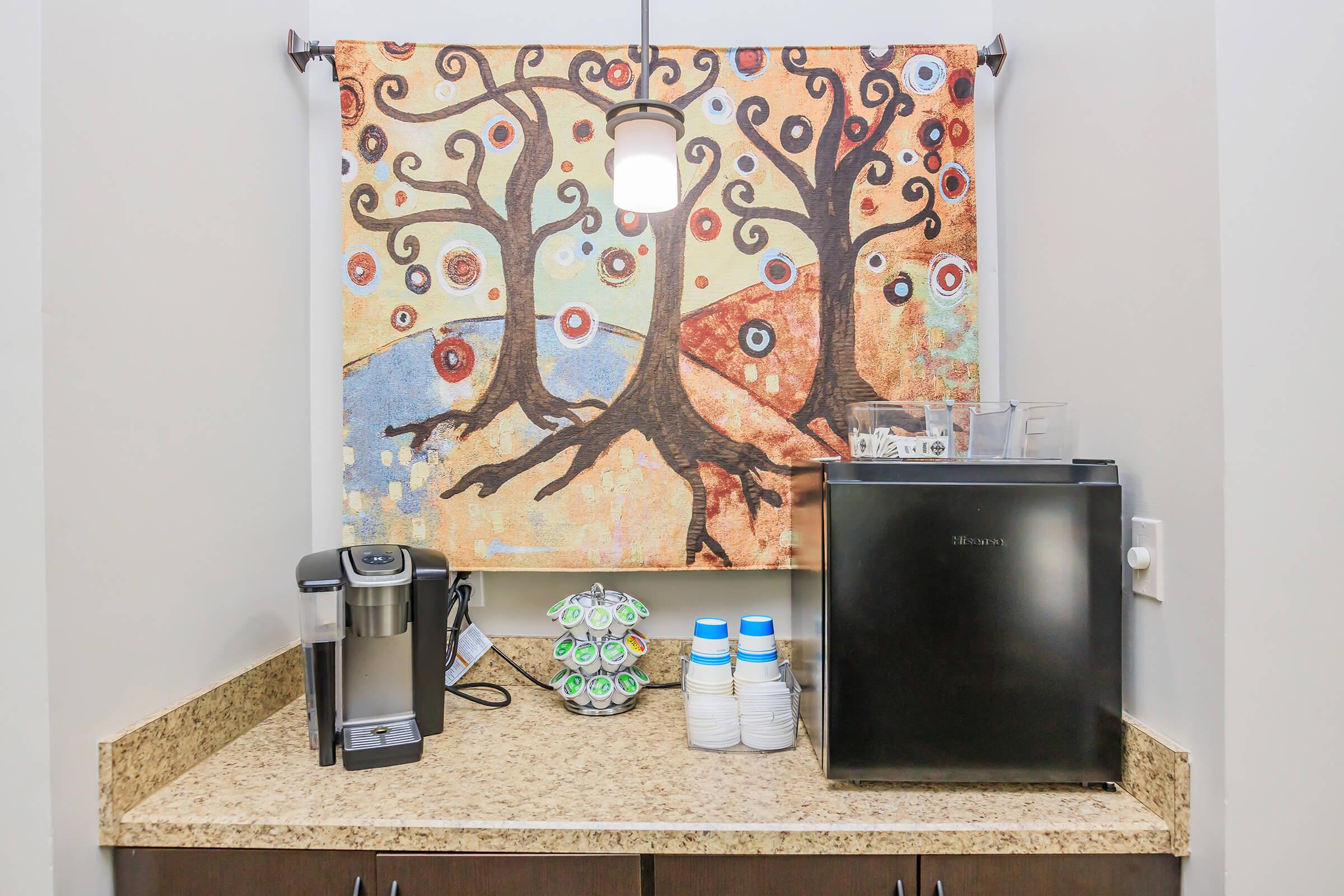 A kitchen counter with a coffee maker, a black mini fridge, cups, and a decorative wall hanging featuring colorful trees on a light background. The artwork adds a vibrant touch to the space.