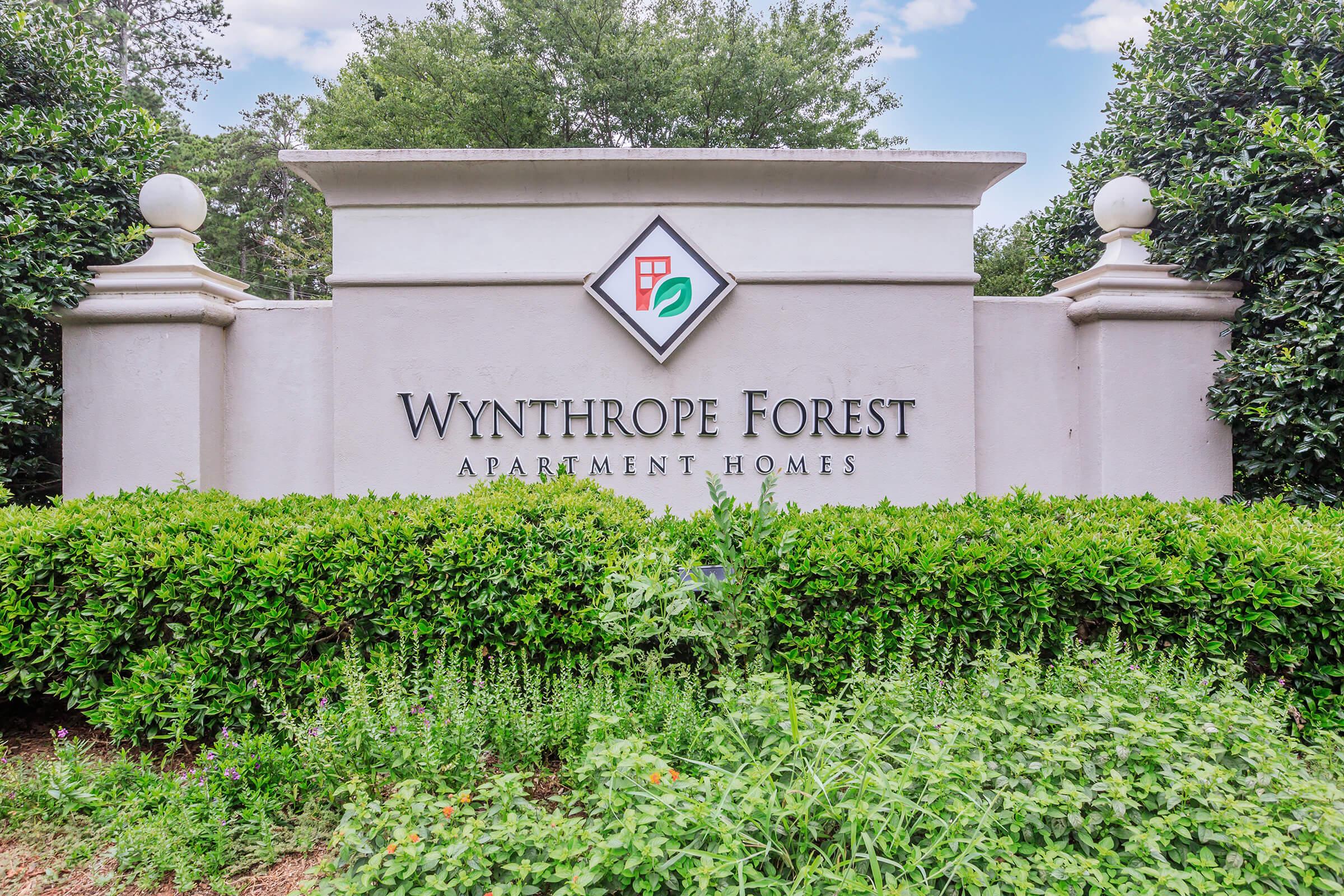 Sign for Wynthrope Forest Apartment Homes, featuring the name prominently displayed with a decorative logo above. Surrounded by manicured greenery and shrubs, with a clear blue sky in the background.