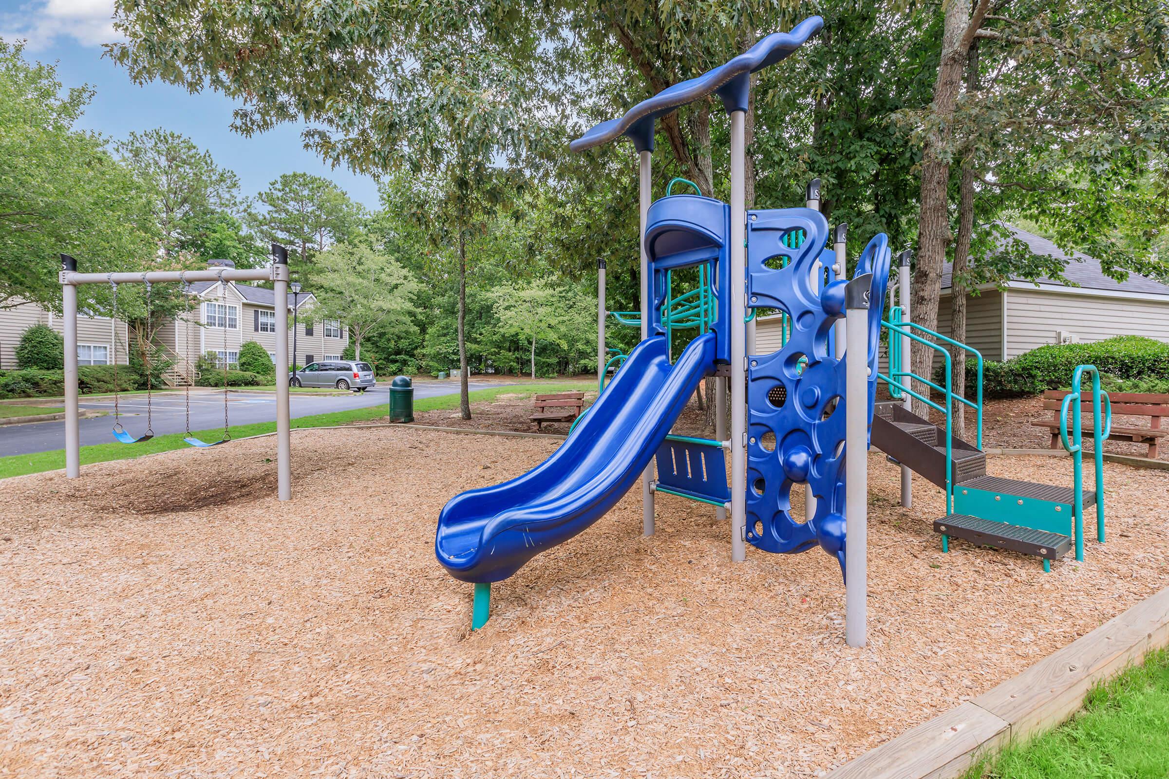 A colorful playground featuring a blue slide, climbing structure, and swings, surrounded by mulch and trees. The playground is situated in a residential area with homes visible in the background.