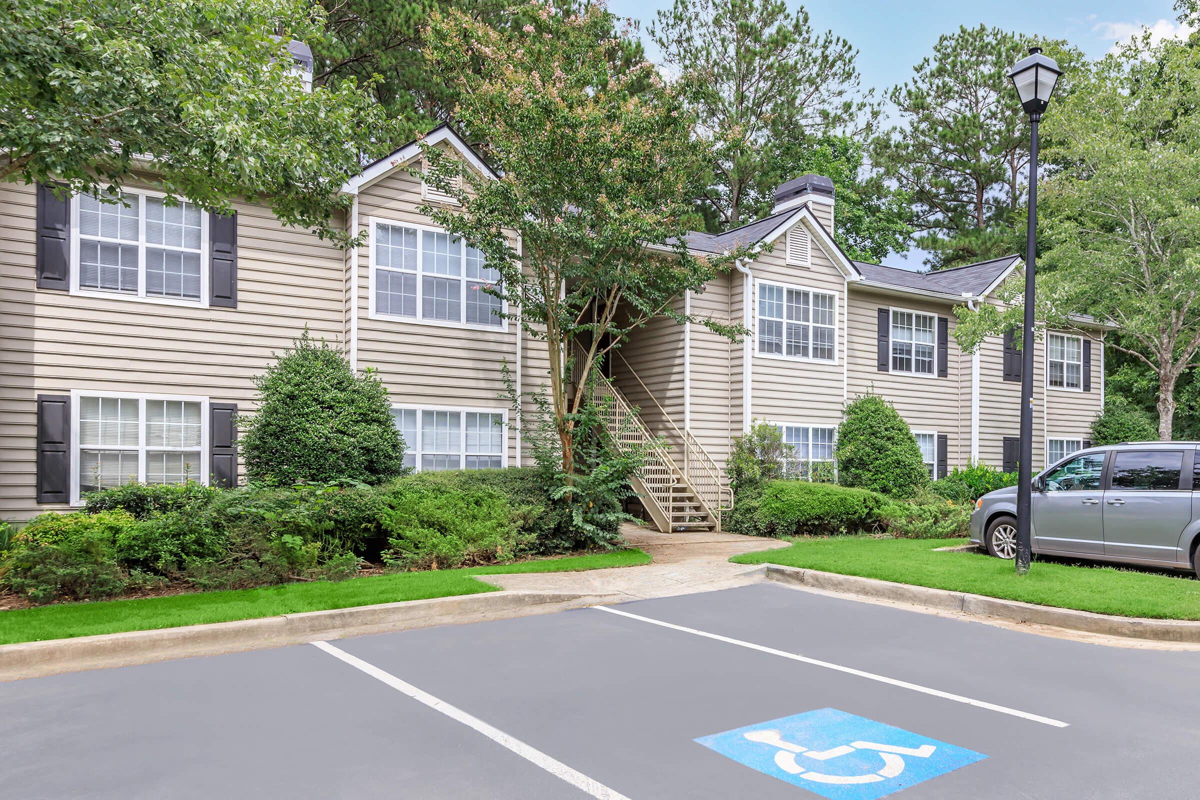 A two-story apartment building with beige siding and white window trim, surrounded by green bushes and trees. There are designated parking spaces in front, including a marked handicapped spot. A silver vehicle is parked nearby, and the area features a clear blue sky.