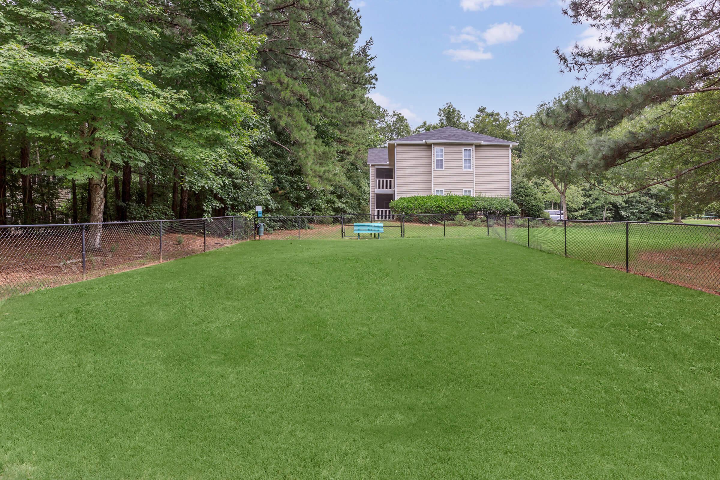A wide view of a green, grassy yard enclosed by a chain-link fence, with a few trees in the background. There is a gray building partially visible in the background, and a small blue bench in the center of the yard, under a clear sky with scattered clouds.