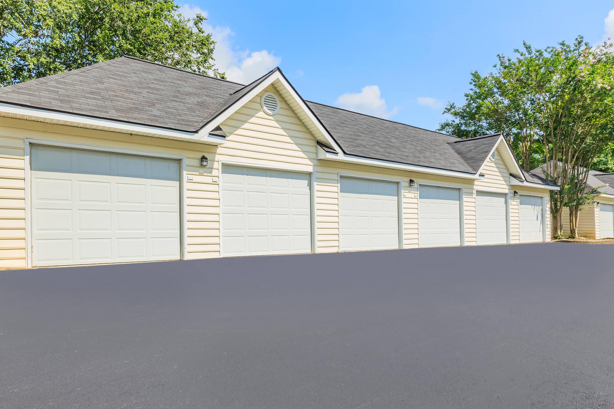 A row of garage doors in a well-maintained parking area, featuring light-colored wooden siding and a clear blue sky in the background. The surface is freshly paved, with trees and greenery visible nearby.