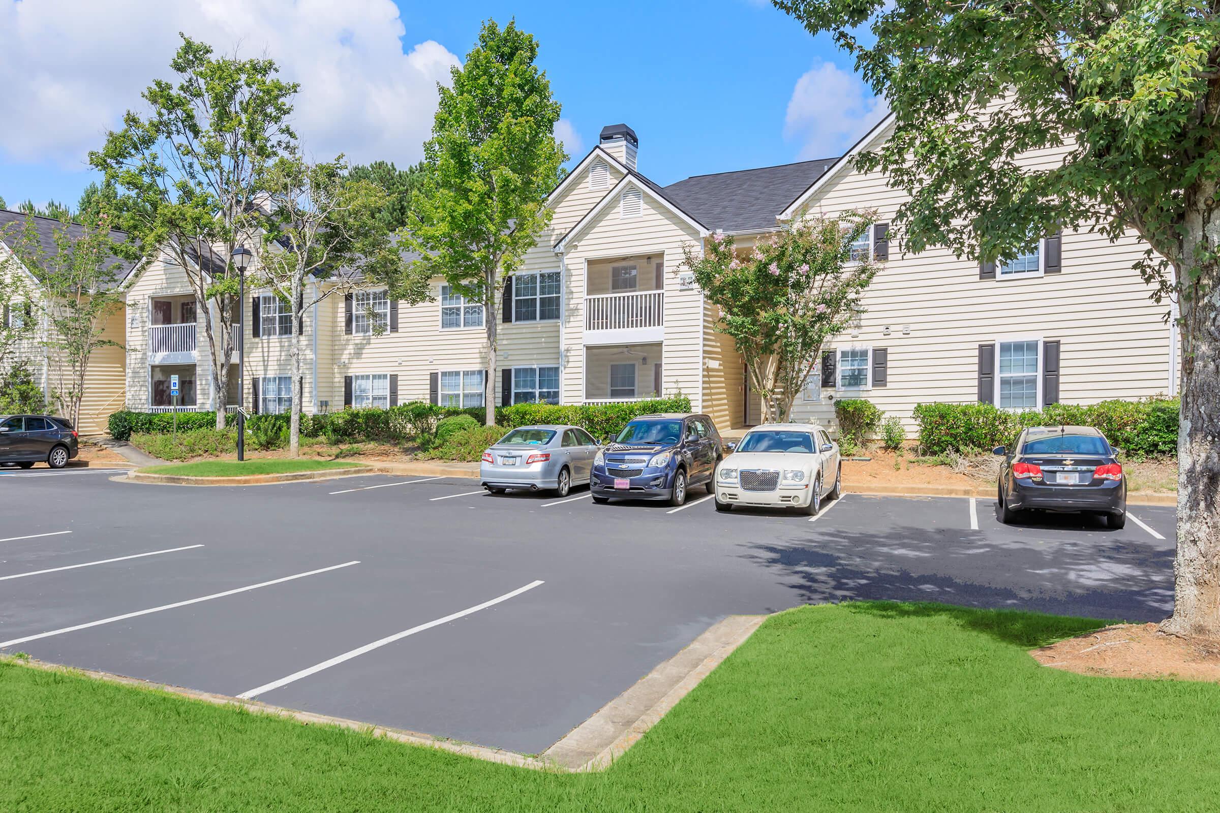 View of a multi-story apartment building with a landscaped area, featuring several parked cars in a lot. The building has a light-colored facade with balconies, green grass, and trees surrounding the premises under a clear blue sky.