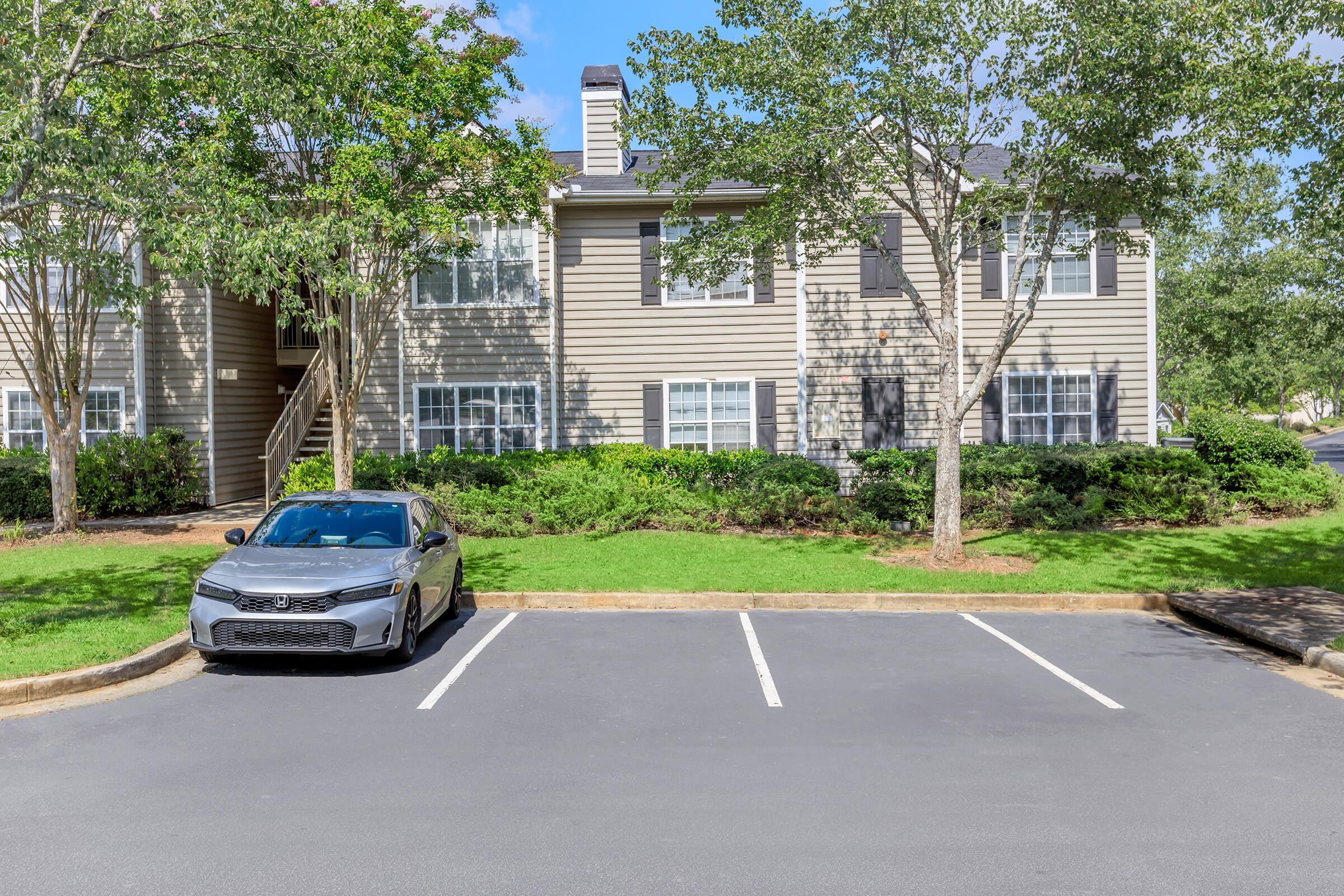 A gray car is parked in front of a two-story apartment building with beige siding and black shutters. The building is surrounded by green grass and trees, and there are several empty parking spaces nearby. The sky is clear and blue, indicating a sunny day.