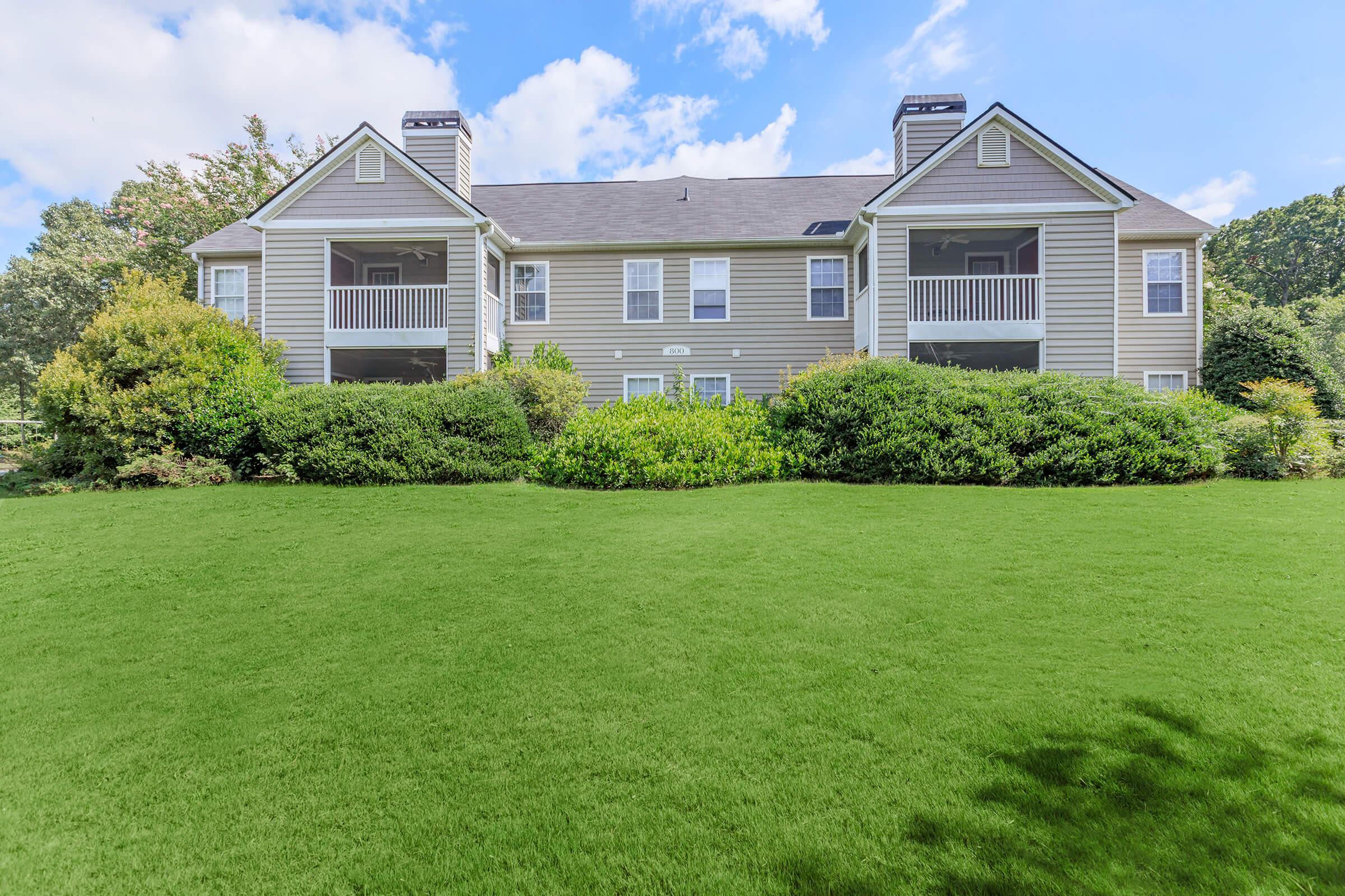 A two-story apartment building with a light gray exterior, featuring multiple balconies and surrounded by lush green landscaping and manicured lawns under a bright blue sky with scattered clouds.