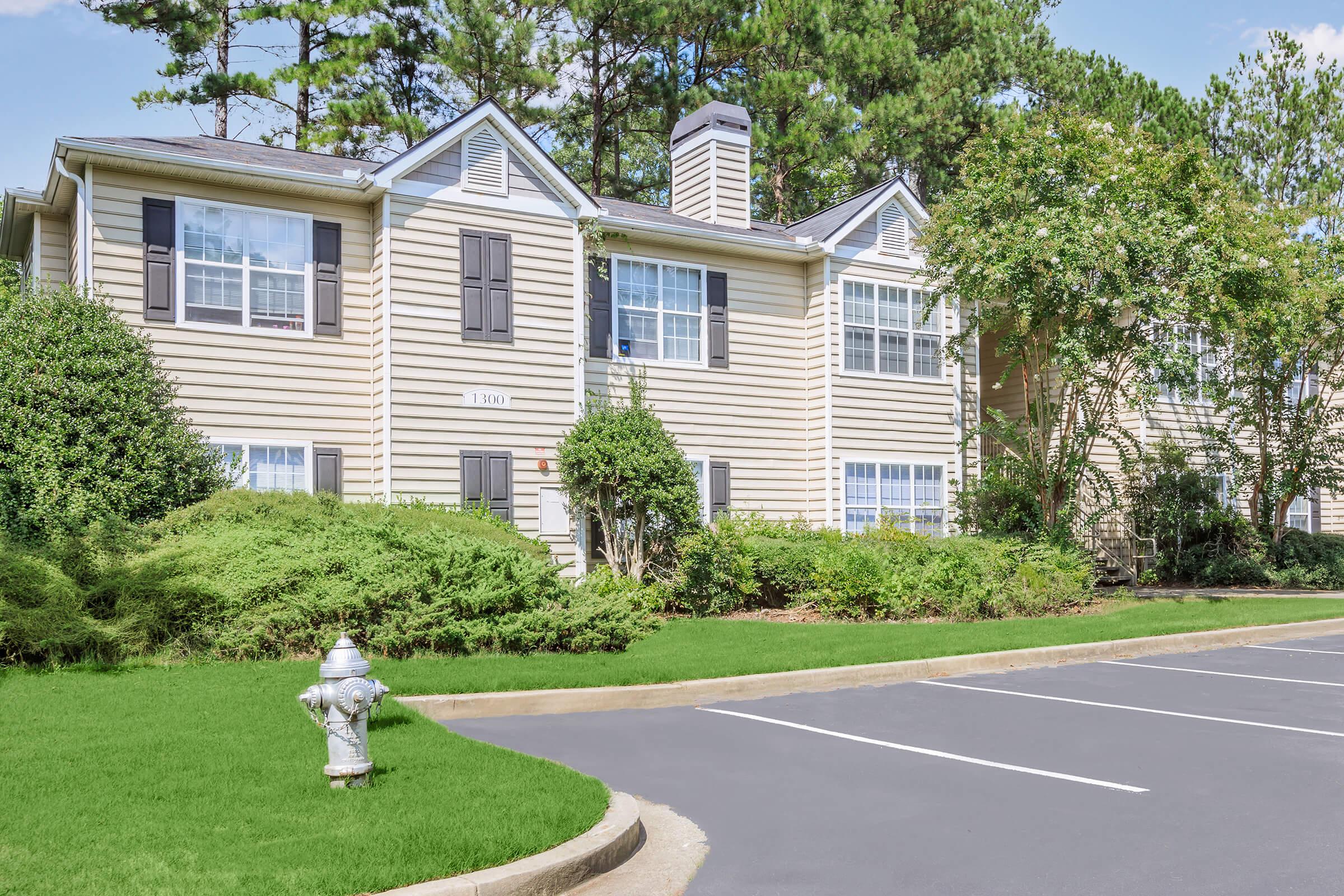 A two-story residential building with beige siding and dark shutters, surrounded by green shrubs and trees. A fire hydrant is visible in the foreground, and a blacktop parking area is nearby. Blue sky with scattered clouds adds to the sunny atmosphere.