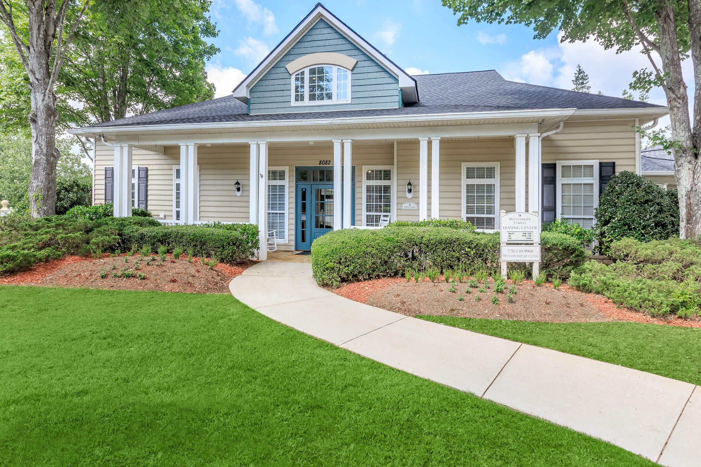 A charming single-story building with light blue and beige siding, featuring a gabled roof and white columns. Lush green grass surrounds the pathway leading to the entrance, which has a light blue door. Neatly trimmed bushes and landscaped flower beds enhance the inviting appearance of the property.