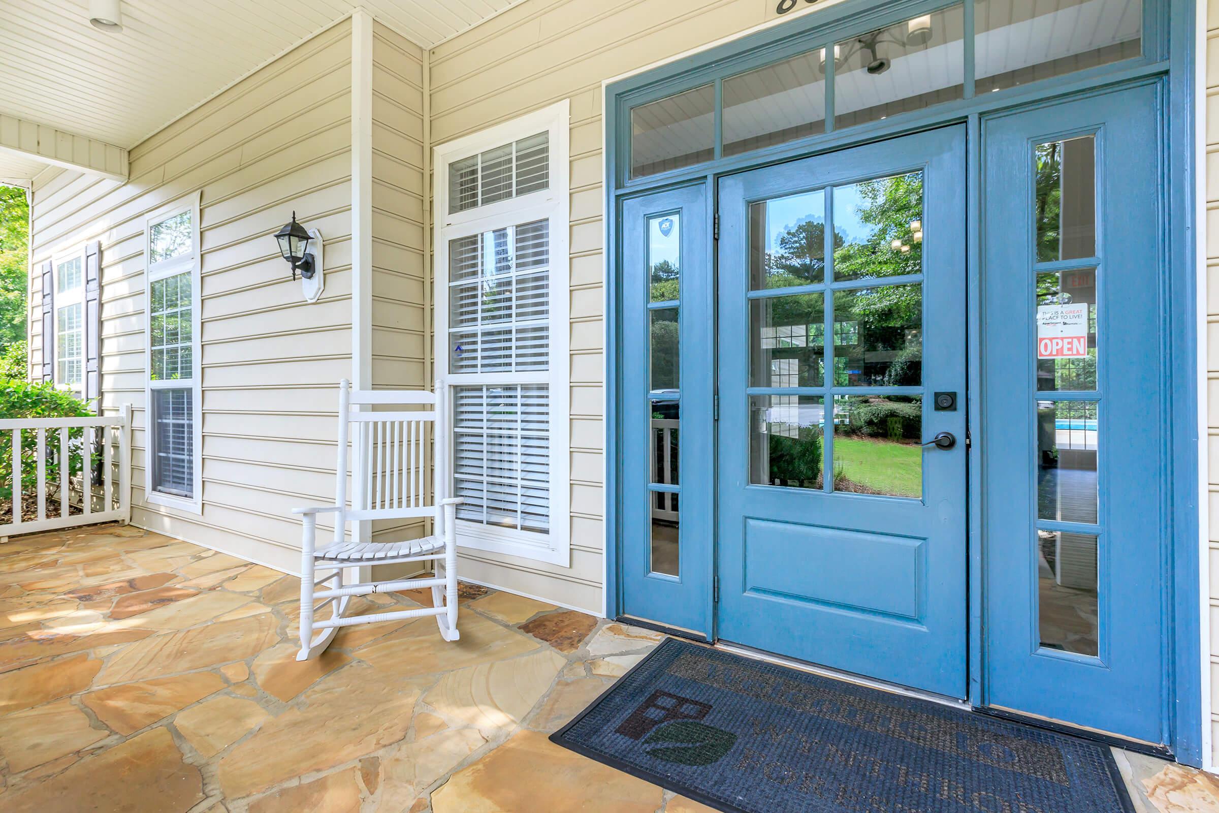 A welcoming porch featuring a blue front door with large windows, a white rocking chair, and a stone pathway. The entrance has a neighborhood charm, with a "OPEN" sign visible through the door, inviting visitors into the home. Lush greenery is seen in the background.