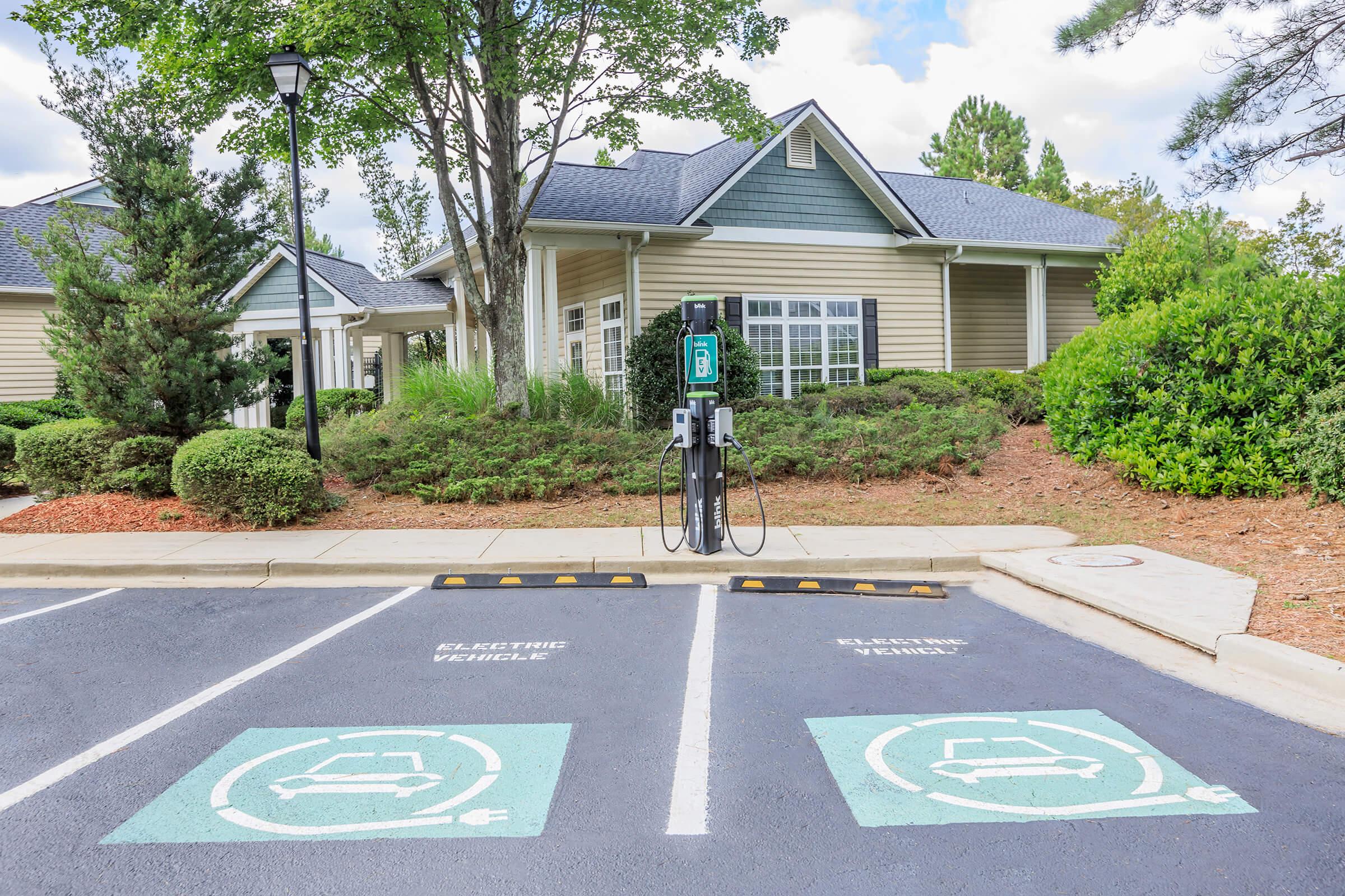 A parking area with designated spots for electric vehicles (EV) next to a charging station. The background features a residential building surrounded by greenery and trees under a clear sky. The EV charging station is prominently displayed in the foreground.