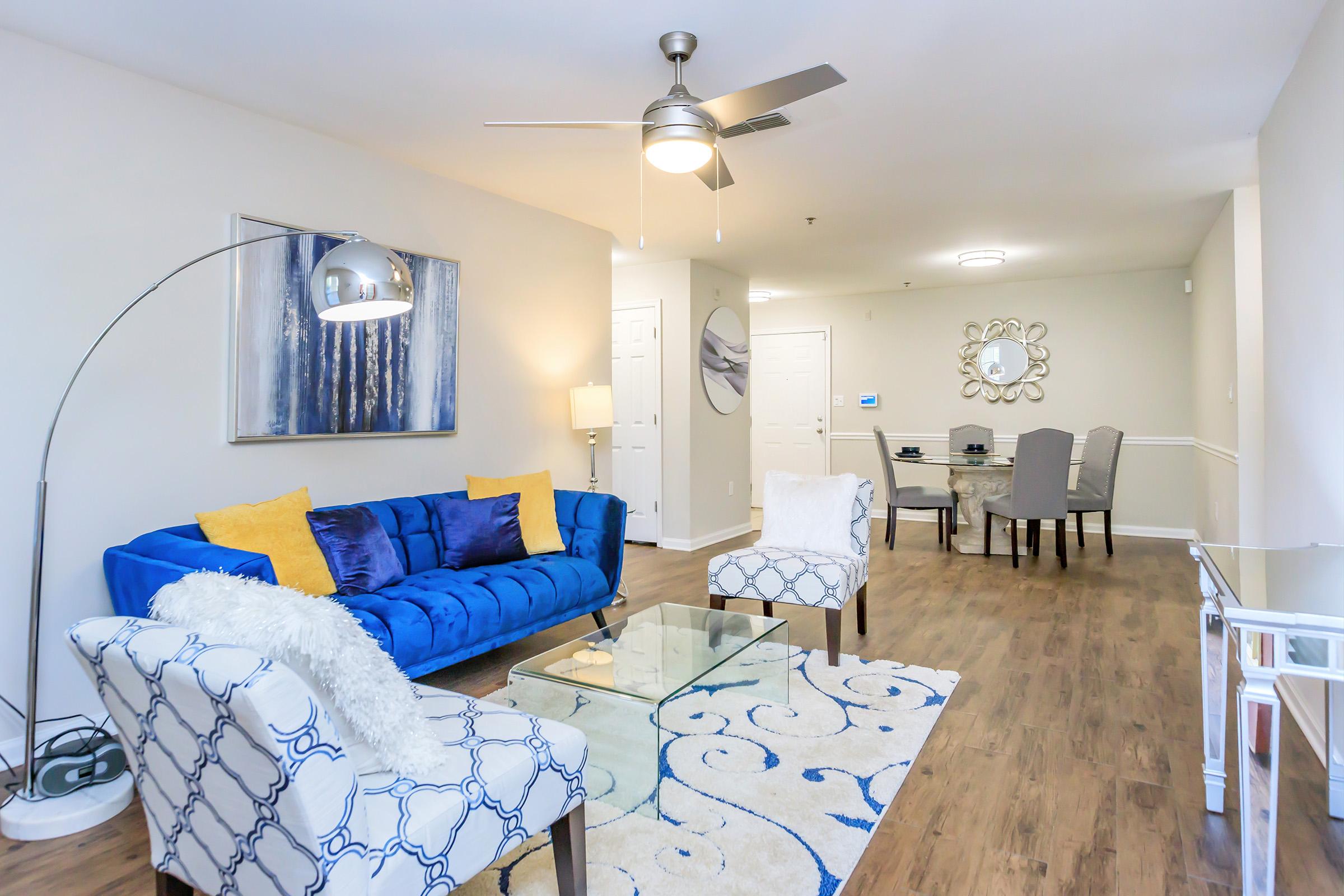 A modern living room featuring a blue sofa with yellow and blue pillows, two patterned armchairs, and a glass coffee table on a decorative rug. A floor lamp and ceiling fan provide lighting. In the background, there's a dining area with light-colored chairs and a mirrored wall decor.