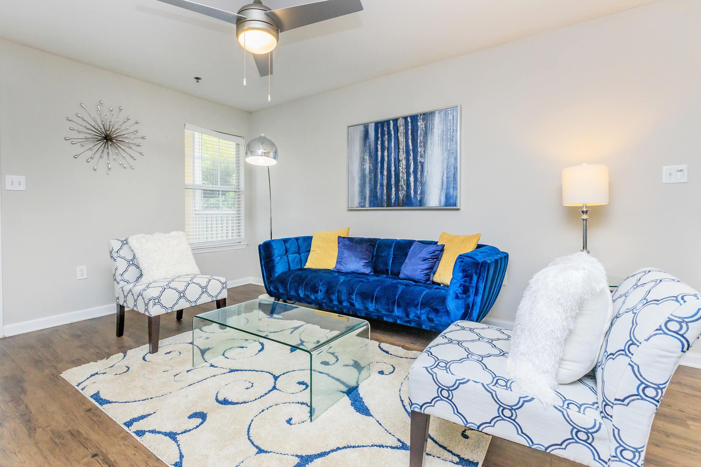 A modern living room featuring a plush blue couch with yellow accent pillows, two patterned chairs, and a glass coffee table. The room has light wood flooring, a large area rug, and soft lighting from a floor lamp. Decorative elements include a wall art piece depicting trees and a starburst wall clock.