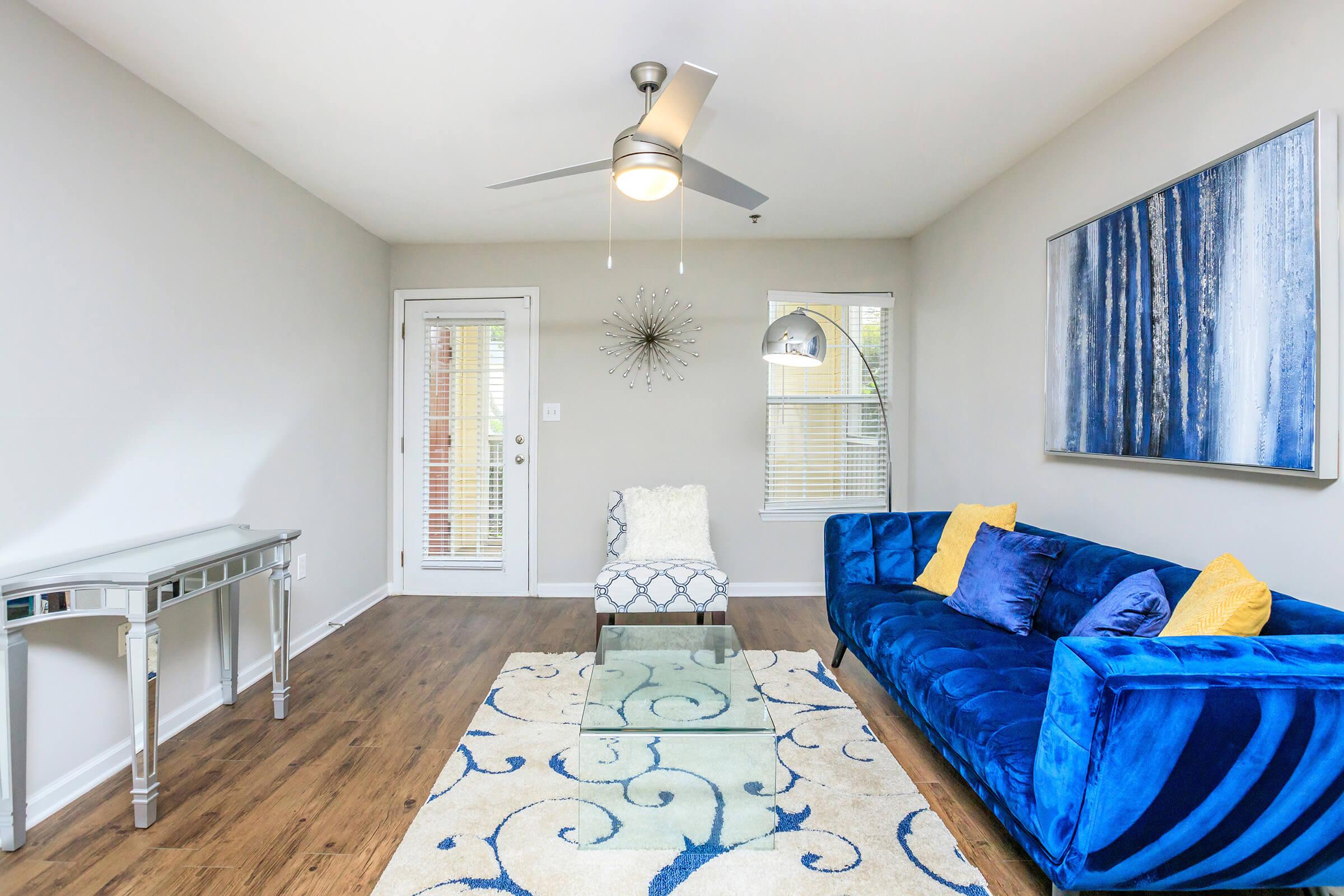 A modern living room featuring a plush blue sofa with yellow cushions, a glass coffee table on a decorative rug, a contemporary chair, and a sleek console table. Bright natural light fills the space through large windows, complemented by a stylish ceiling fan and abstract wall art.