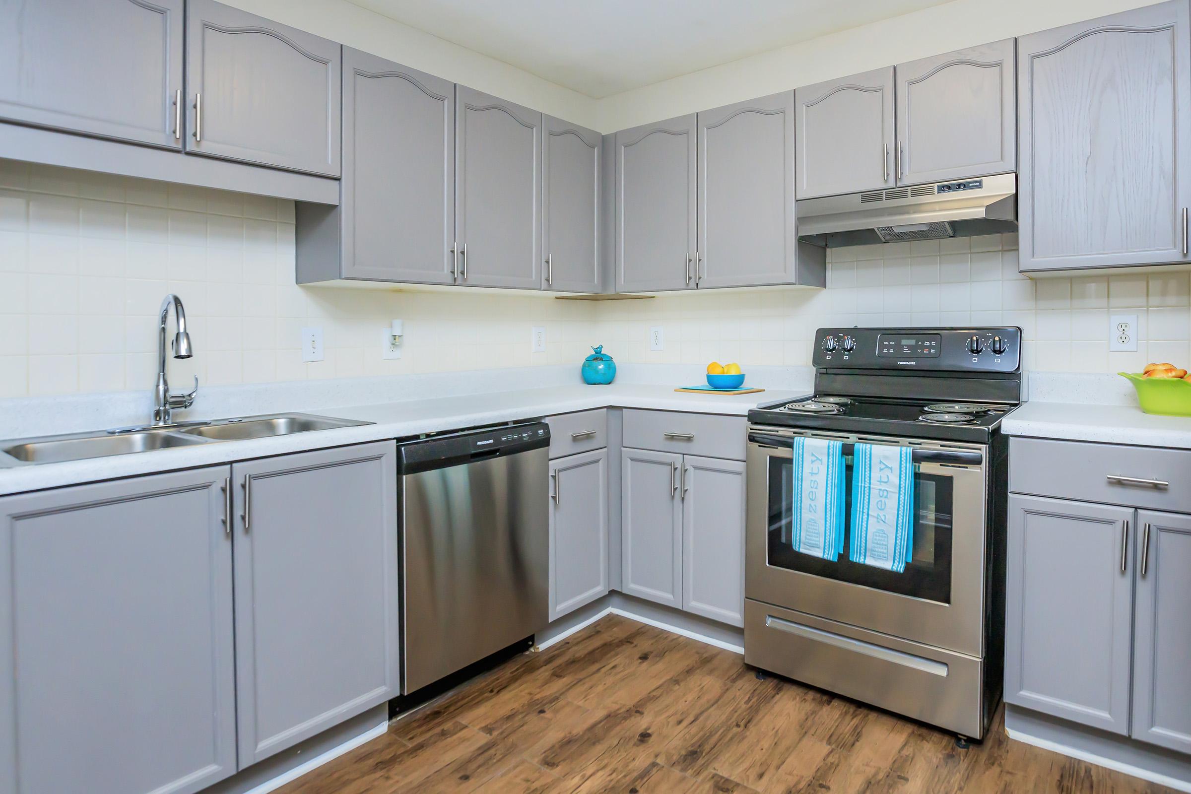 Modern kitchen featuring light gray cabinets, a stainless steel sink, dishwasher, and oven. Light-colored countertops and backsplash complement the cabinetry. The kitchen includes some colorful decorative items, such as a blue bowl and a green bowl with fruit. The flooring is warm wood laminate.