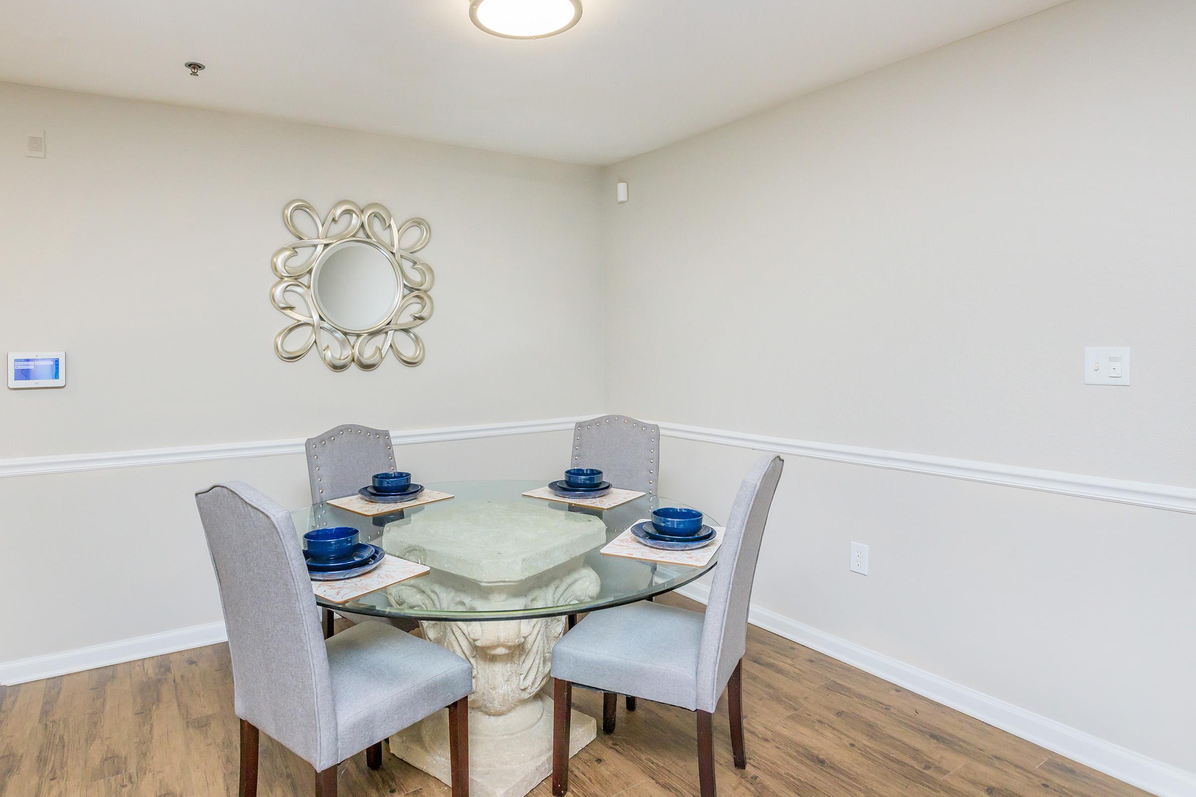 A modern dining area featuring a round glass table with a stone base, surrounded by four light gray upholstered chairs. The table is set with blue plates and white placemats. A decorative mirror is hung on the wall, and the flooring is wooden, enhancing the contemporary feel of the space.
