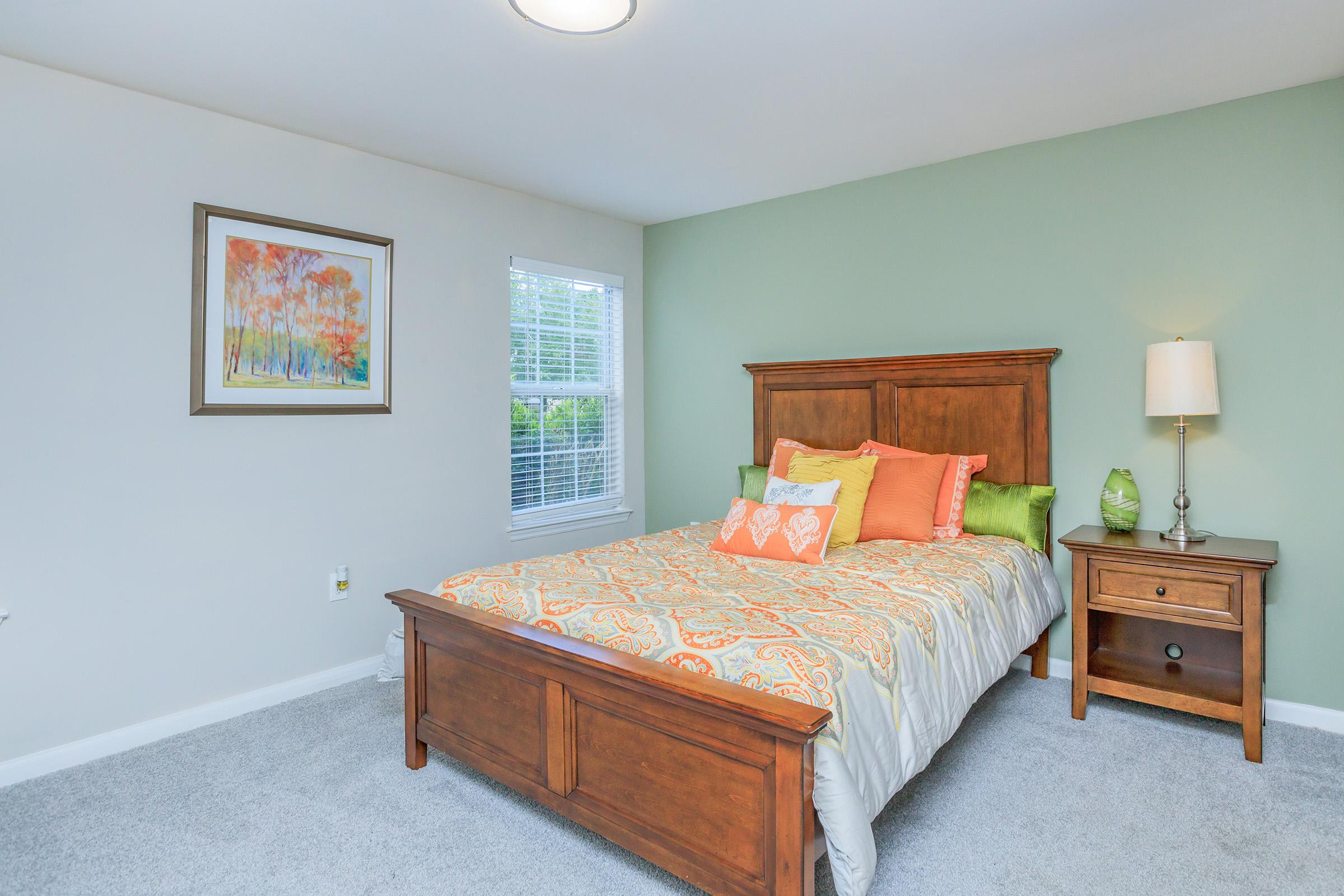Cozy bedroom featuring a wooden bed frame with a colorful patterned comforter and decorative pillows. A bedside table with a lamp and a green decor item complements the space. The walls are painted a soft green, and there’s a framed artwork depicting trees in vibrant autumn colors. Natural light streams in through a window.