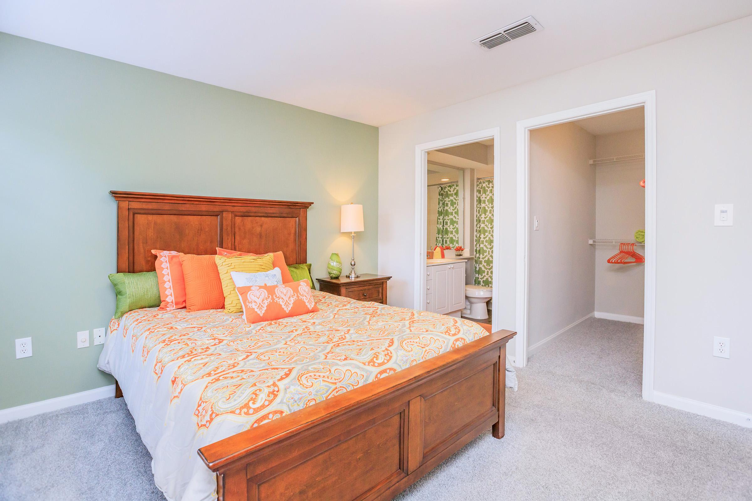 A cozy bedroom featuring a wooden bed frame with an orange and green patterned comforter. The space includes decorative pillows, a bedside lamp, and a small dresser. A doorway leads to a bathroom with a shower curtain visible. The room has light gray carpet and soft green walls.