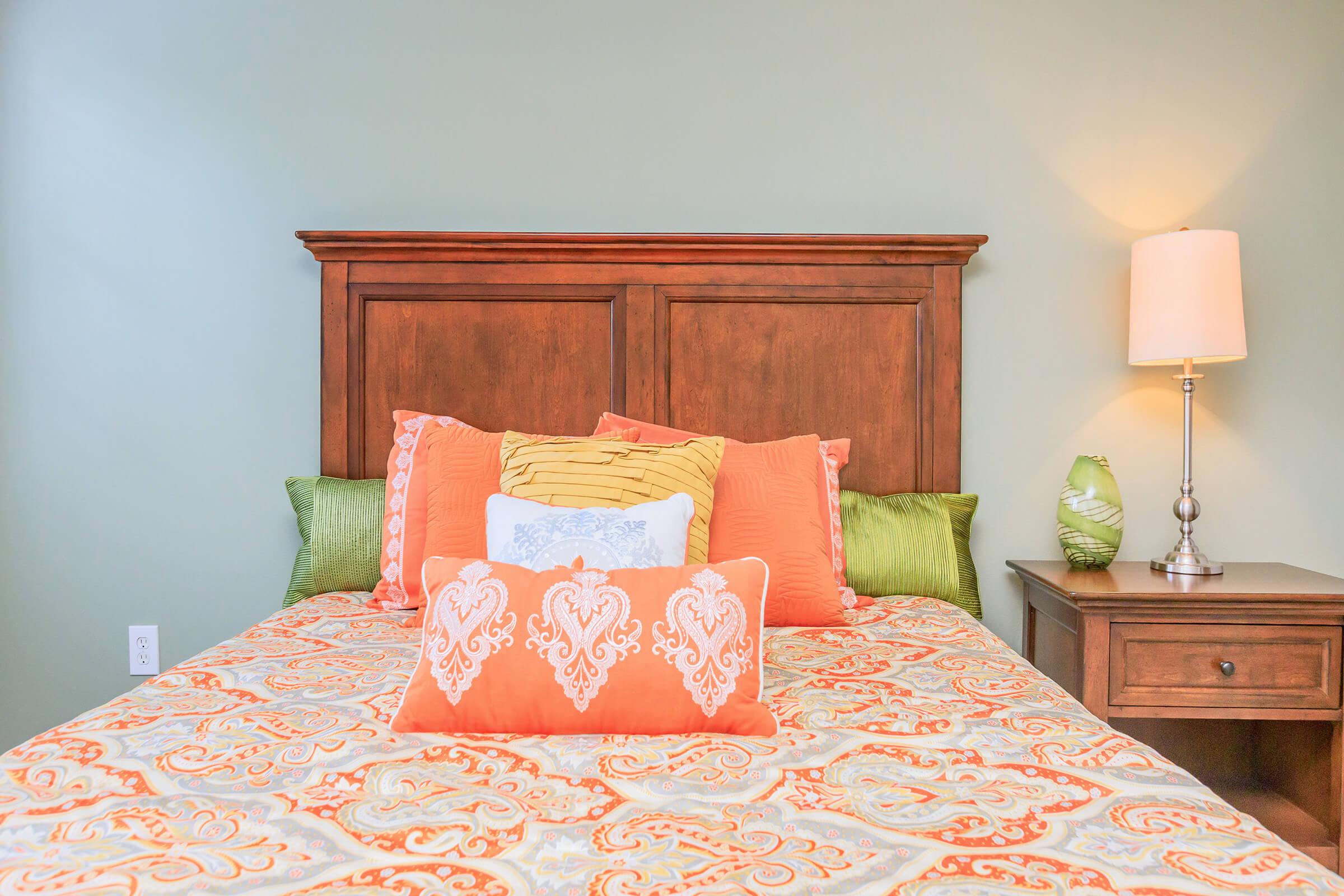 A cozy bedroom featuring a wooden bed frame with vibrant orange and green pillows. The bedding is adorned with an ornate pattern, and there is a decorative pillow at the center. A bedside table with a lamp is on the right, and a green accent piece is next to the lamp against a soft, muted wall color.
