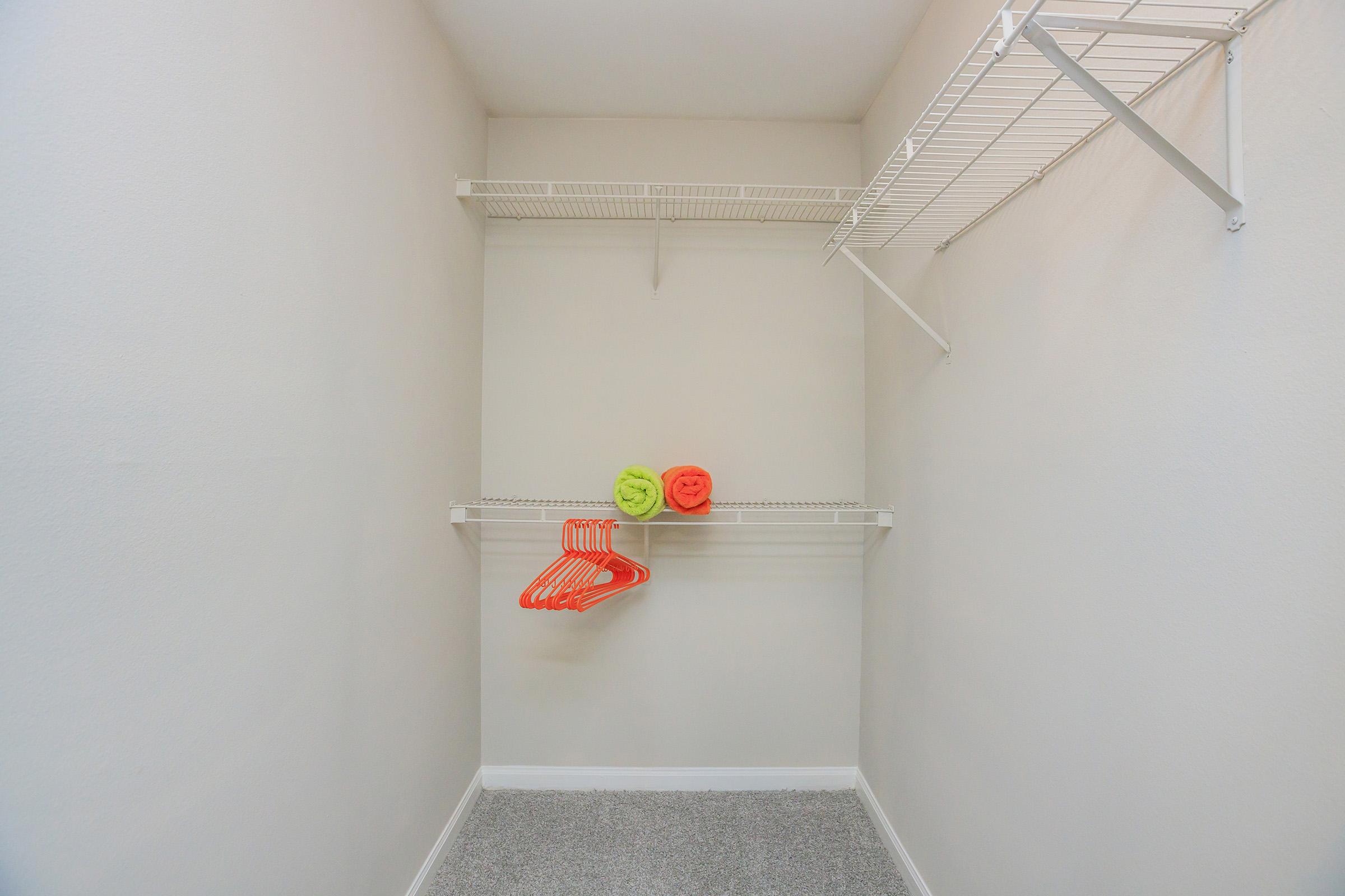 Empty closet interior featuring white wire shelves. Two brightly colored towels, one green and one orange, are neatly placed on the bottom shelf. An additional set of orange plastic hangers hangs from the upper shelf. The walls and flooring are light-colored, creating a clean and spacious look.