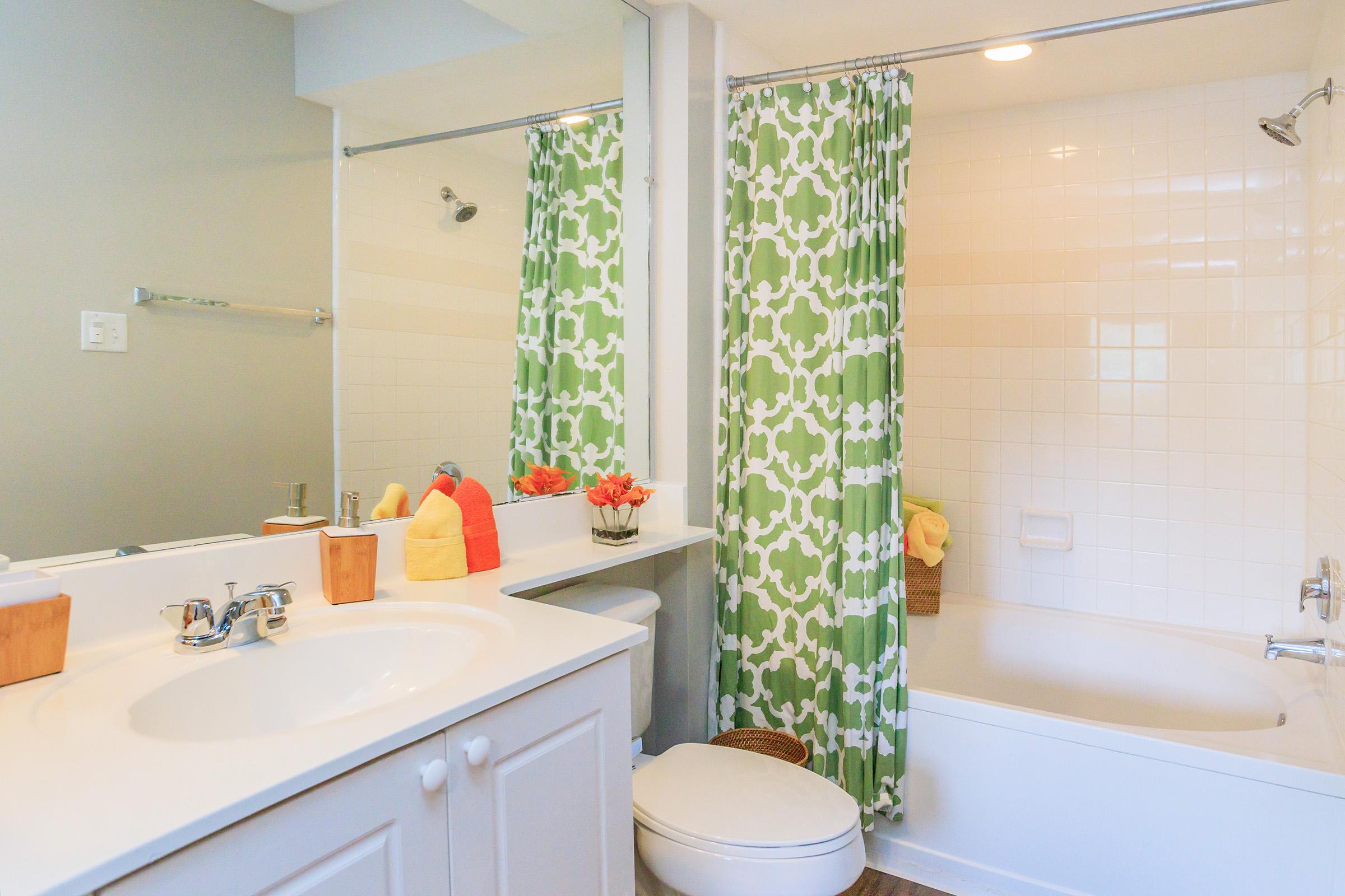 Brightly decorated bathroom featuring a white sink with a mirror, a bathtub with a shower, and a green and white patterned shower curtain. Decorative items include colorful towels and a floral arrangement, complemented by a clean and modern aesthetic.