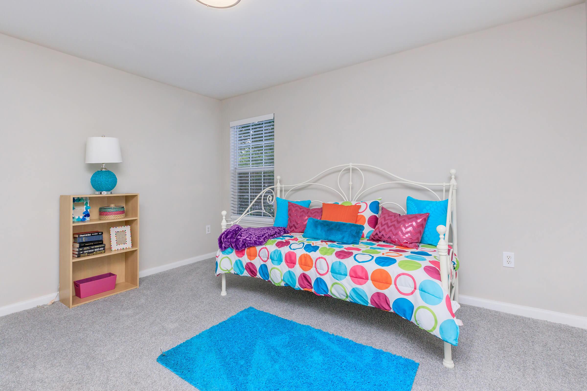 A brightly decorated bedroom featuring a daybed with a colorful polka dot cover, accented by vibrant cushions in pink and blue. A plush blue rug is on the gray carpeted floor, and a wooden bookshelf with books and decorative items is against the wall. A lamp on a side table adds to the cozy ambiance.