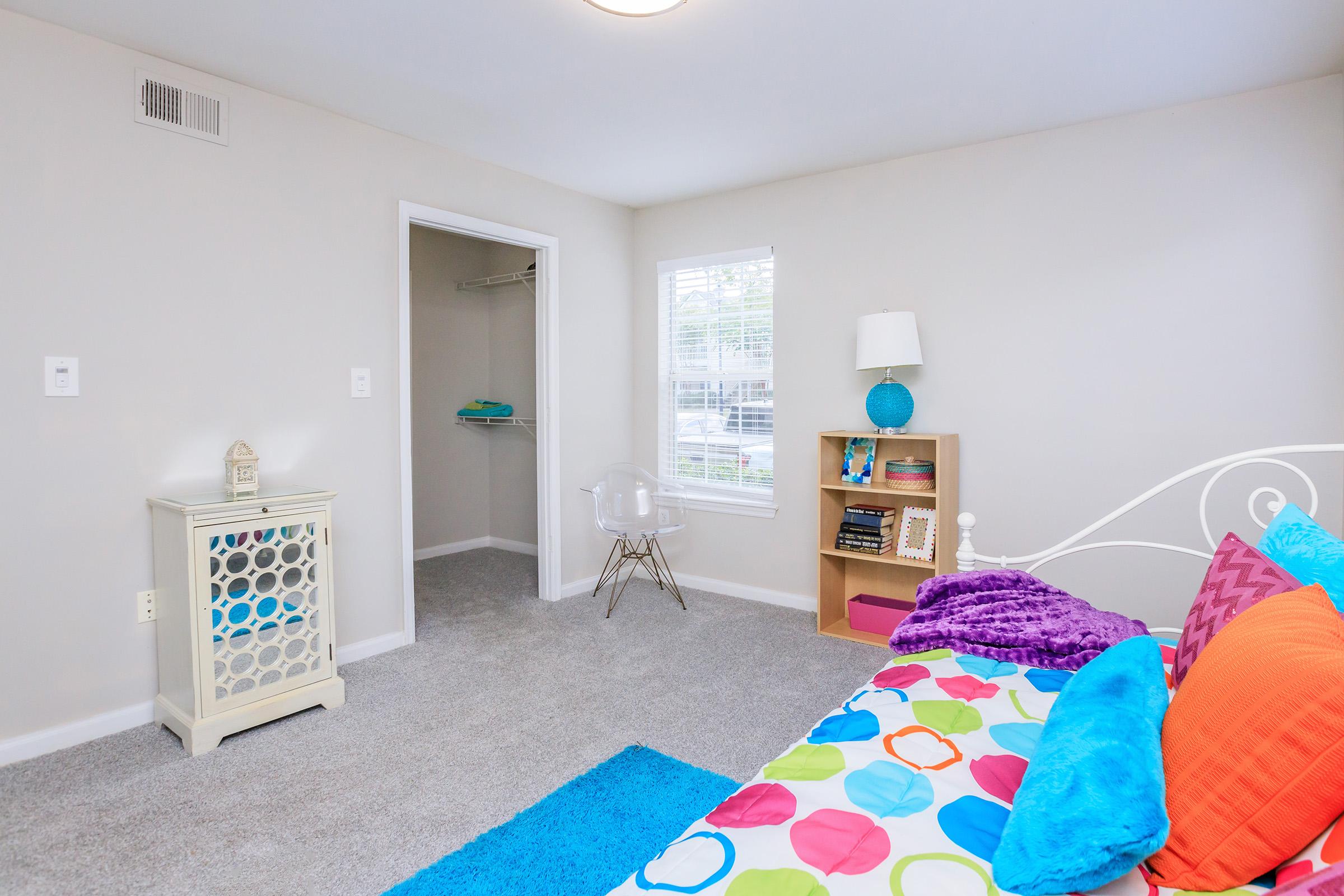 A brightly decorated bedroom featuring a colorful polka dot bedspread, a white bed with decorative pillows, a blue area rug, a small bookshelf with books, and a lamp on a nightstand. There's a clear chair and a closet visible in the background, with natural light coming through the window.