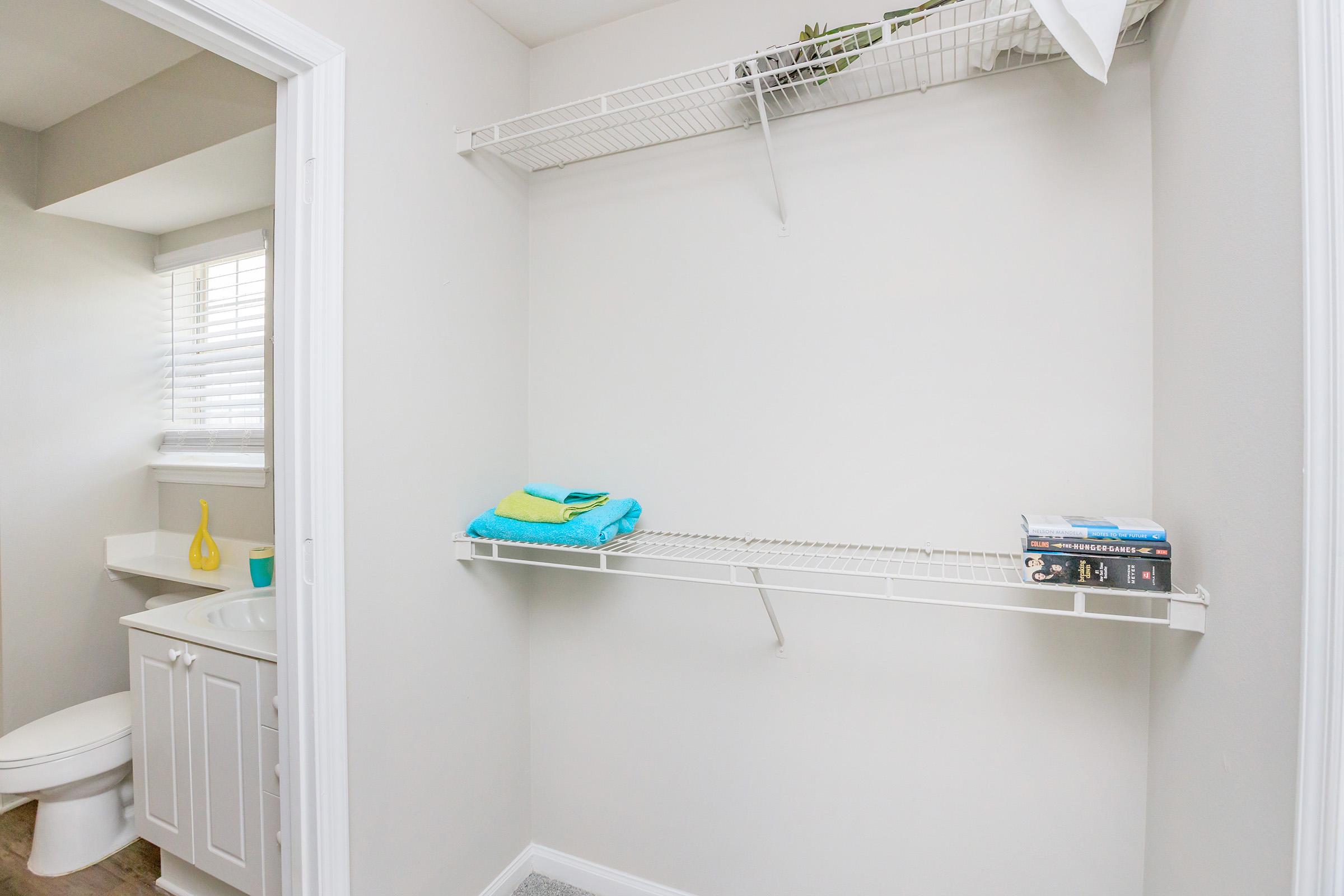 A small bathroom space featuring a white wall, a shelf with neatly folded towels, and some small books. A toilet is visible in the corner, and a sink area is partially shown. Natural light comes in from a nearby window, creating a bright and airy atmosphere.