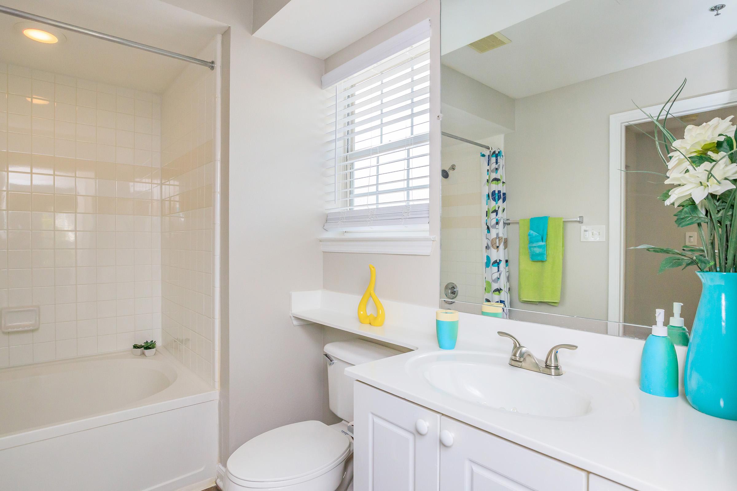 A bright and modern bathroom featuring a white tub and shower, a white vanity with a sink, and a large mirror. There is a vibrant blue vase with flowers on the counter, colorful towels hanging, and natural light coming through a window with blinds. The decor is clean and inviting.