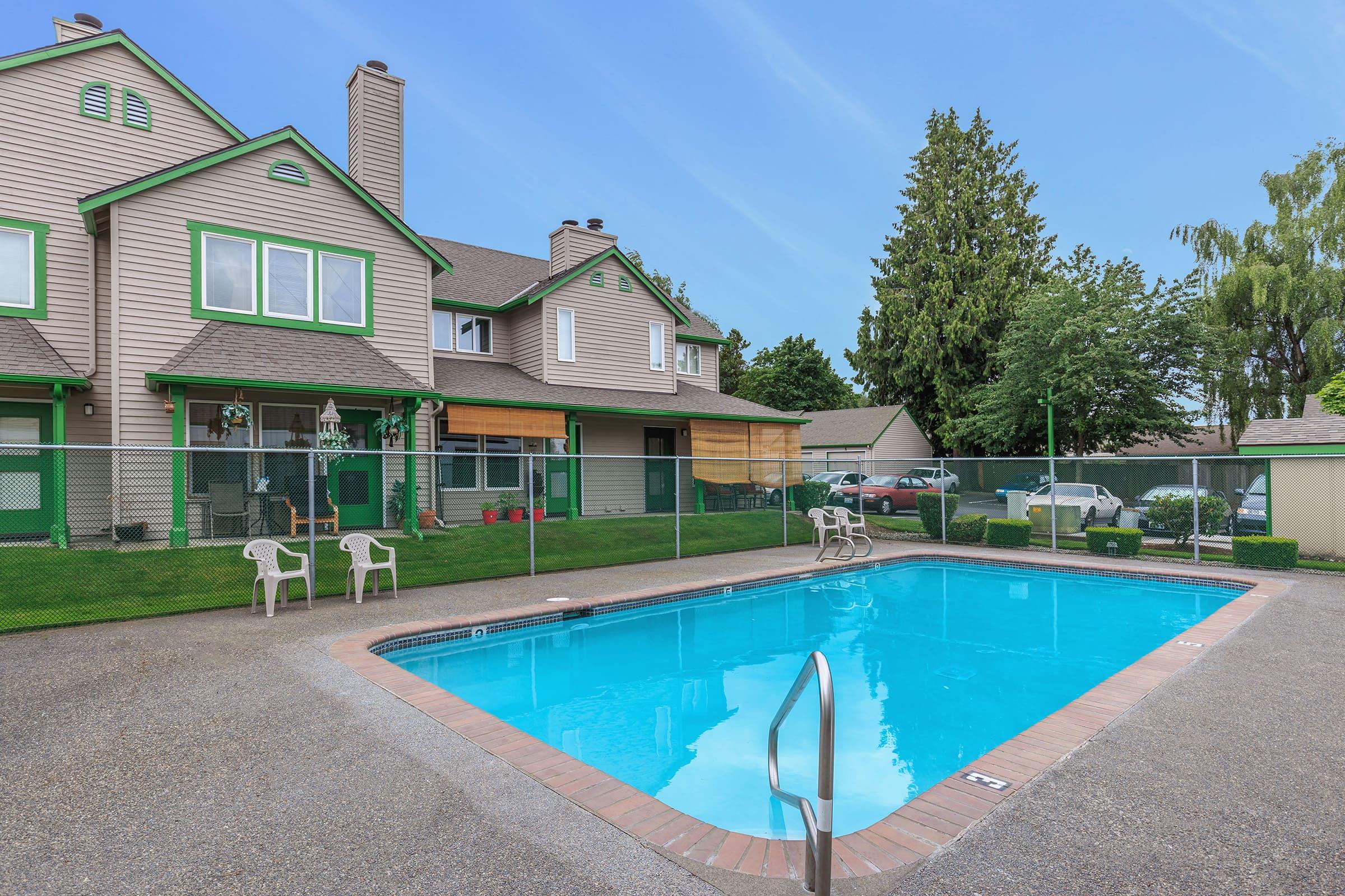 A clean, inviting outdoor swimming pool surrounded by a fenced area. There are lounge chairs positioned around the pool, and a well-maintained grassy area nearby. I
