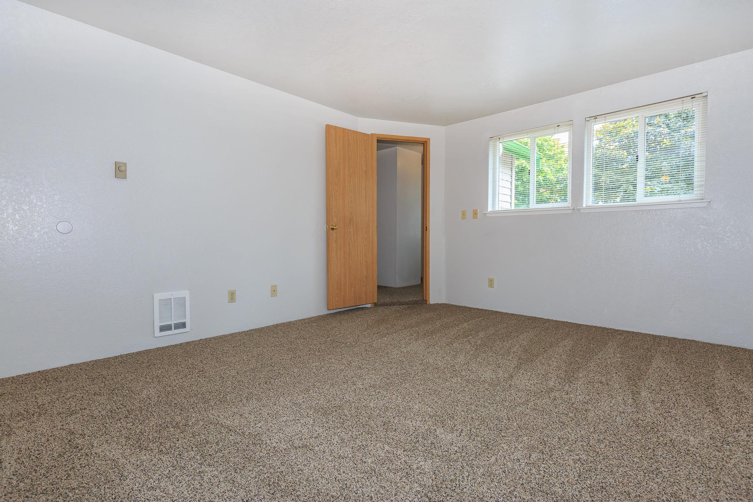 Empty room with light-colored walls and beige carpet. There is a closed wooden door leading to another space and two windows allowing natural light.