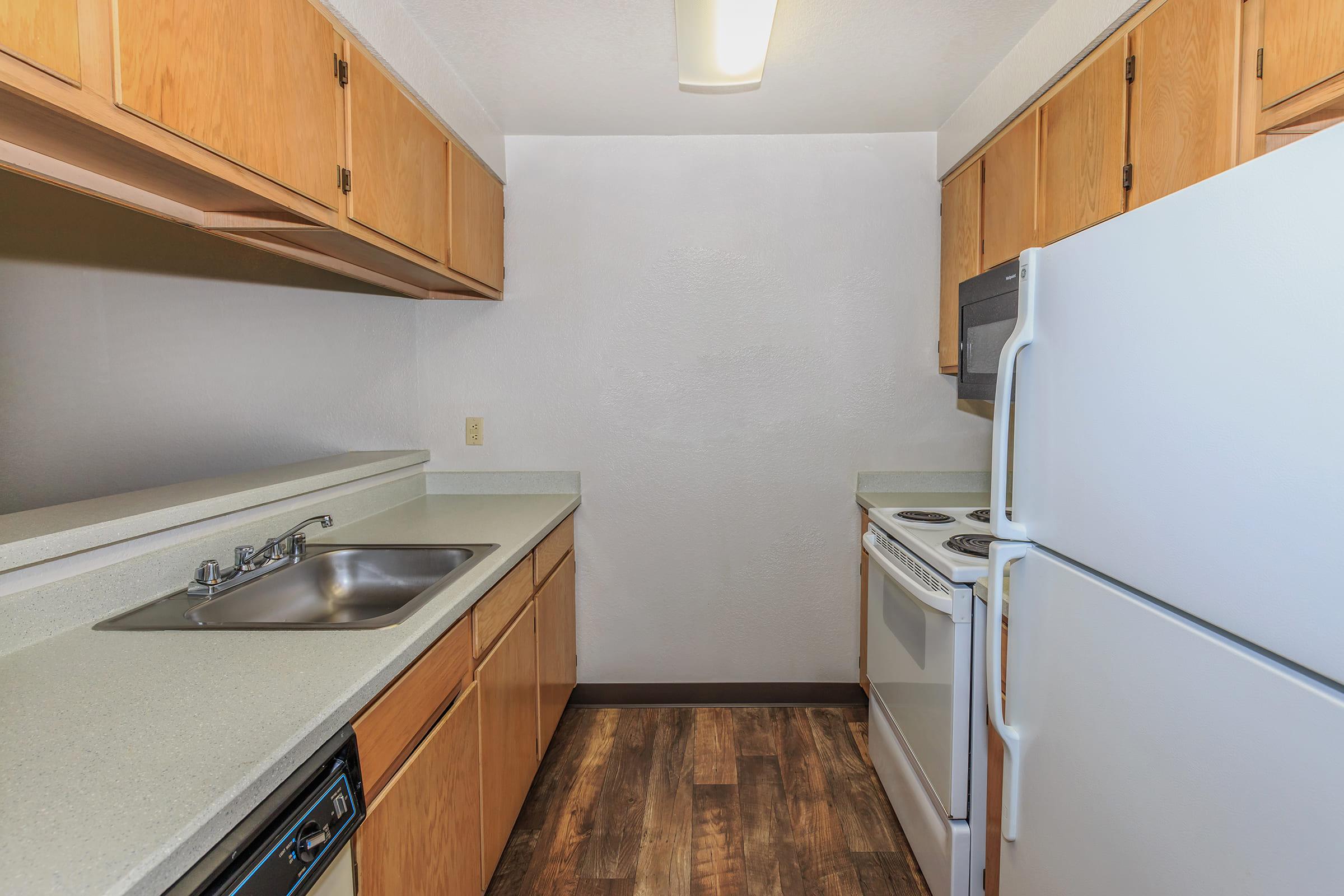 A simple kitchen with light wood cabinets, a stainless steel sink, a white stove, and a refrigerator. The countertops are gray, and the floor features dark wood-like laminate.