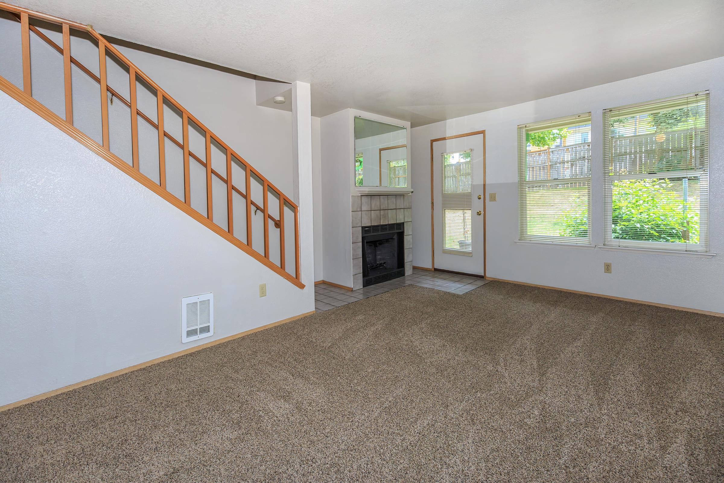 Spacious living area featuring tan carpeting, a staircase with wooden railing, a cozy fireplace, large windows allowing natural light, and a doorway leading outside.