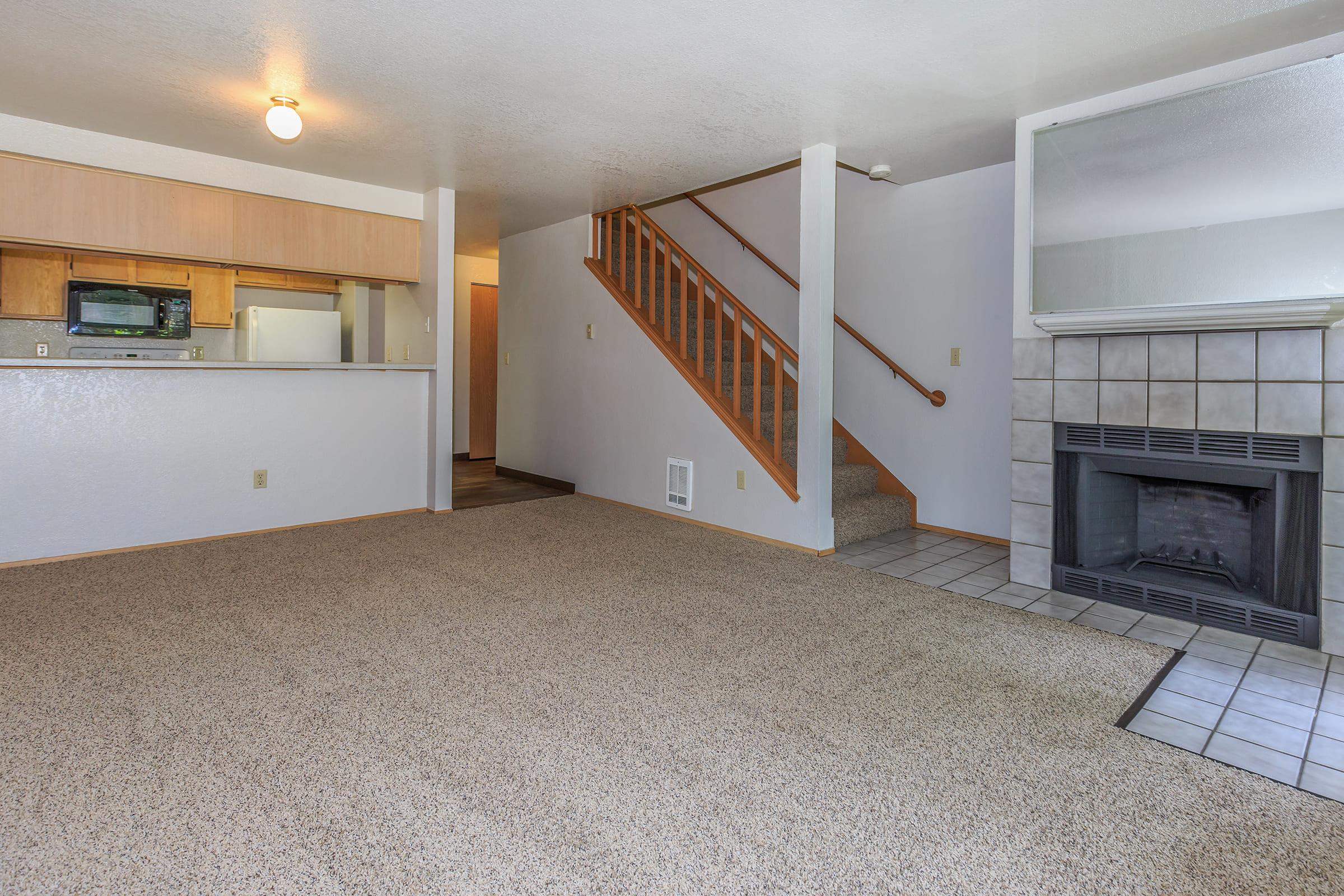 Interior view of a cozy living room with light-colored carpet, a fireplace, and wooden stairs leading to an upper level.