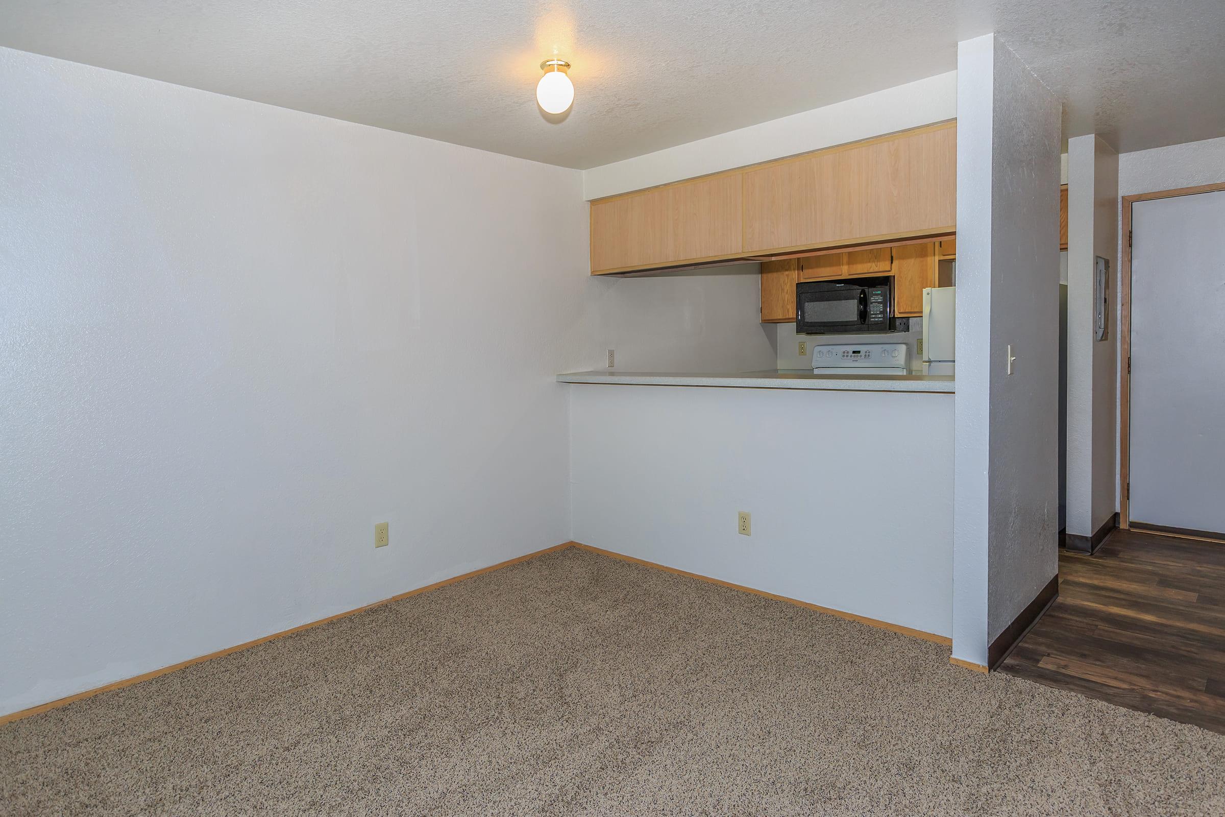 A spacious, unfurnished room with light beige carpeting and white walls. In the background, there's a kitchen area with wooden cabinets, a microwave, and a stove.
