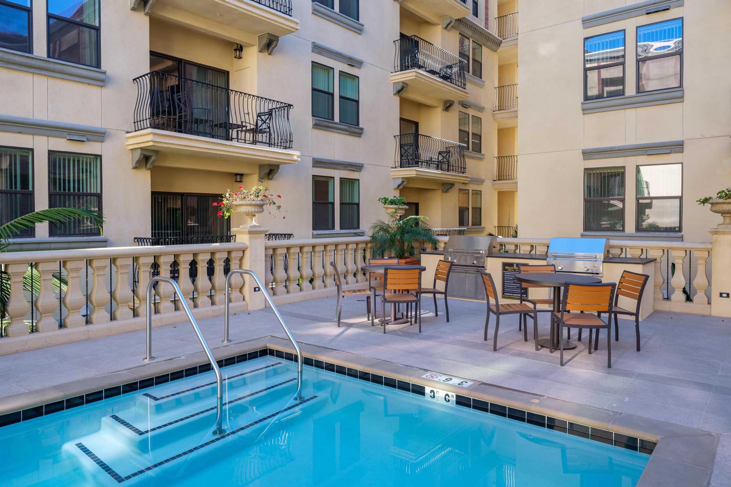 A serene apartment complex courtyard featuring a swimming pool with stairs, surrounded by a patio area with outdoor dining furniture and a grill. The building has multiple levels with balconies, and potted plants add a touch of greenery to the space.