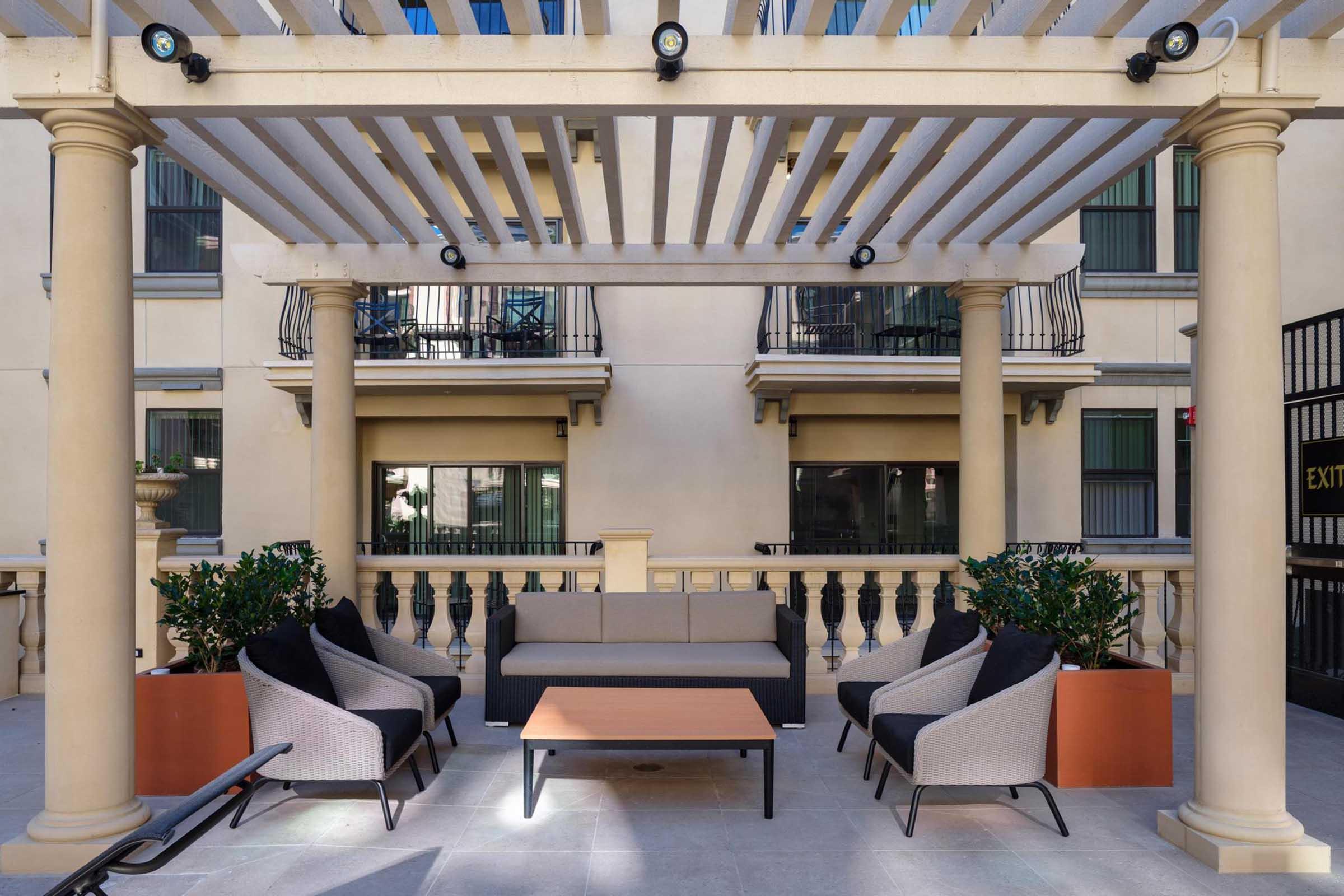 Outdoor lounge area with a modern design featuring a pergola overhead supported by columns. There are four comfortable chairs arranged around a coffee table, with potted plants nearby. The background shows a building with balconies and windows, creating a cozy and inviting atmosphere.